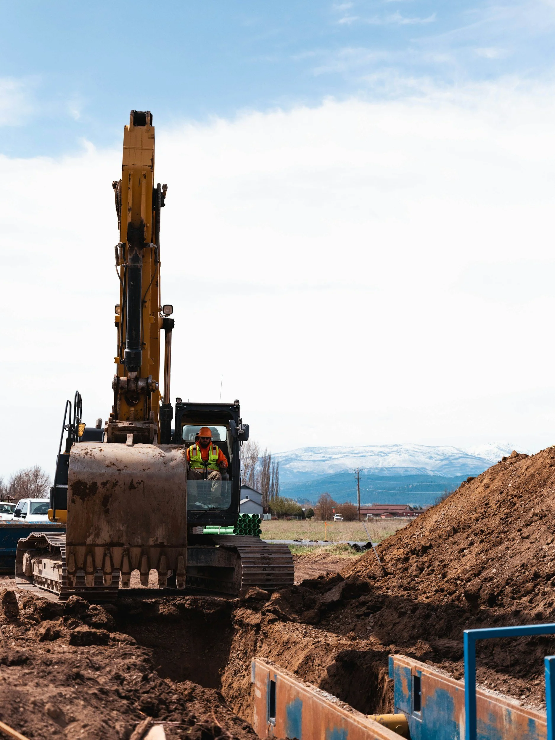 Construction worker operating a yellow excavator in a dirt field, with mountains in the background.