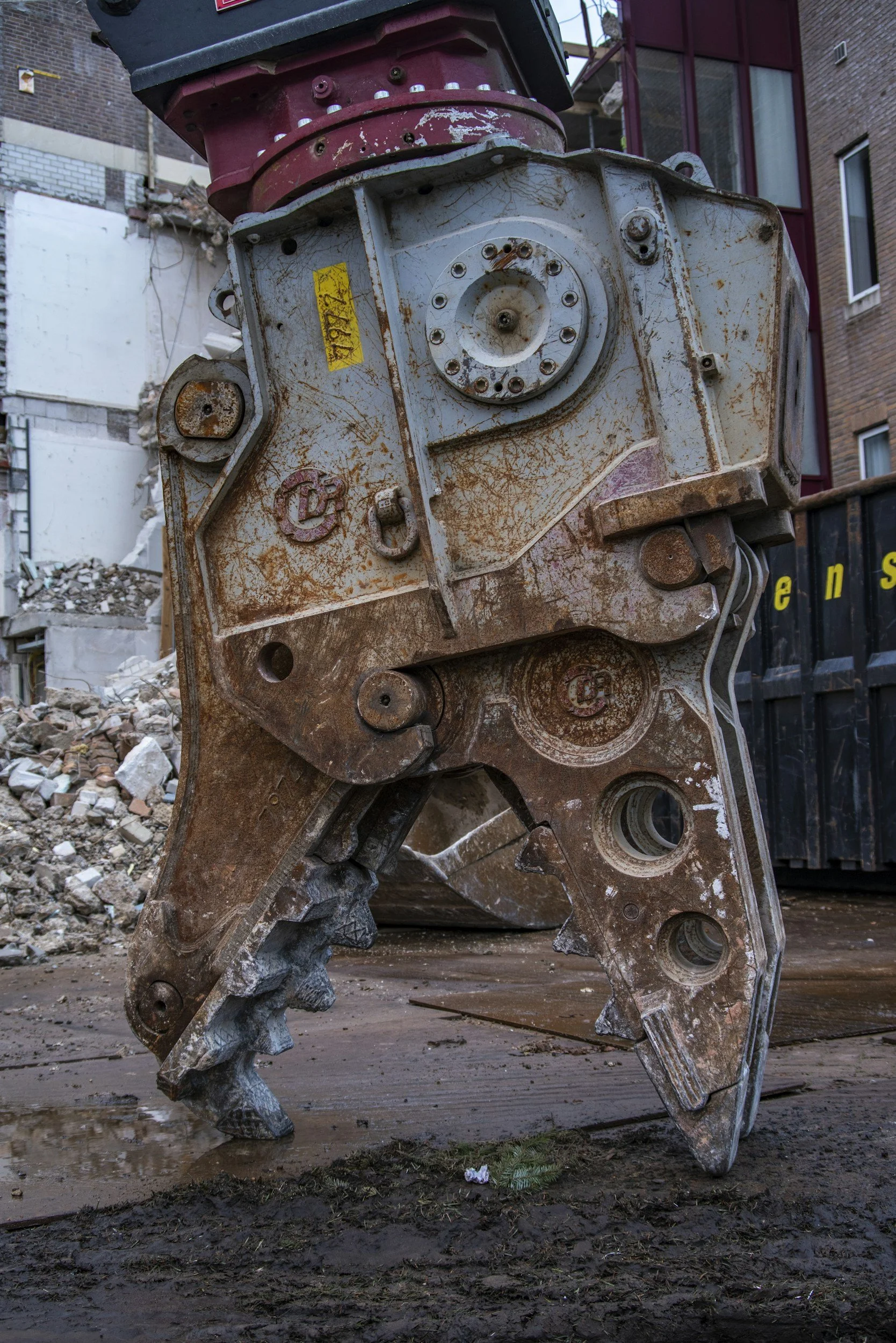 Close-up of a large, rusty construction machine component with metal gears and bolts, on a construction site with debris and bricks in the background.