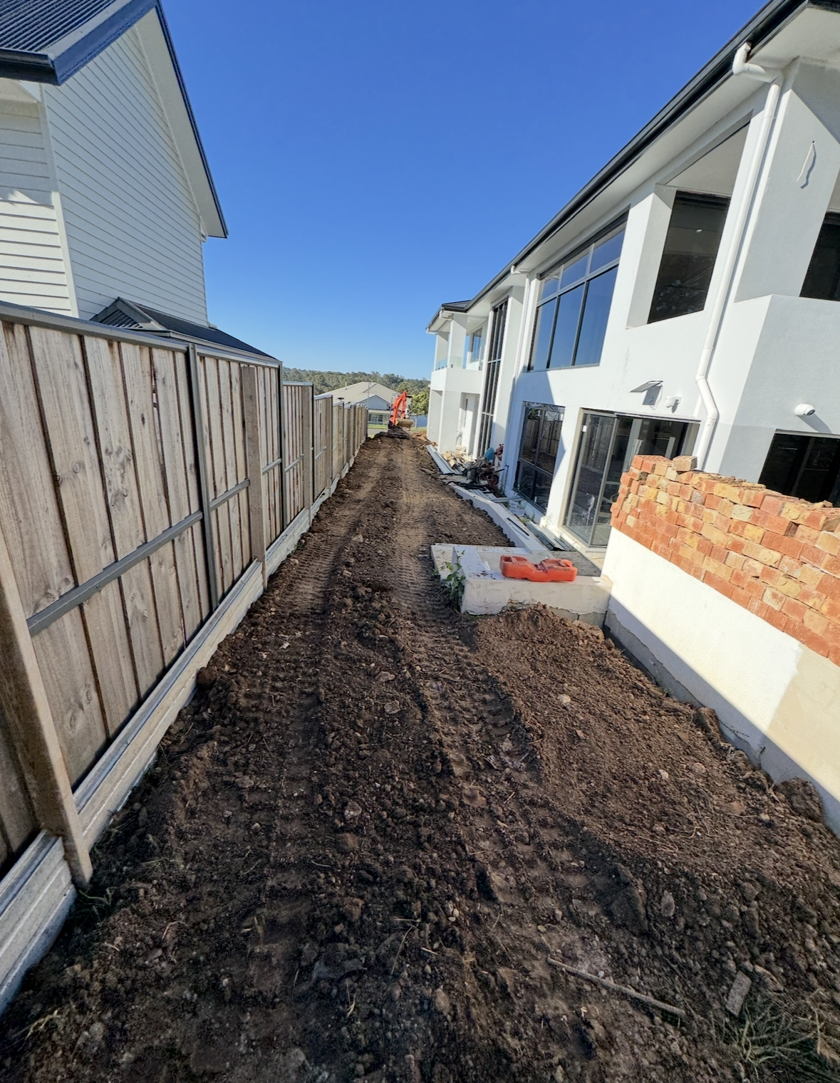A construction site showing a dirt pathway between a wooden fence on the left and a modern house on the right with large windows. The area appears to be under development with construction materials and equipment visible, and the sky is clear and blu