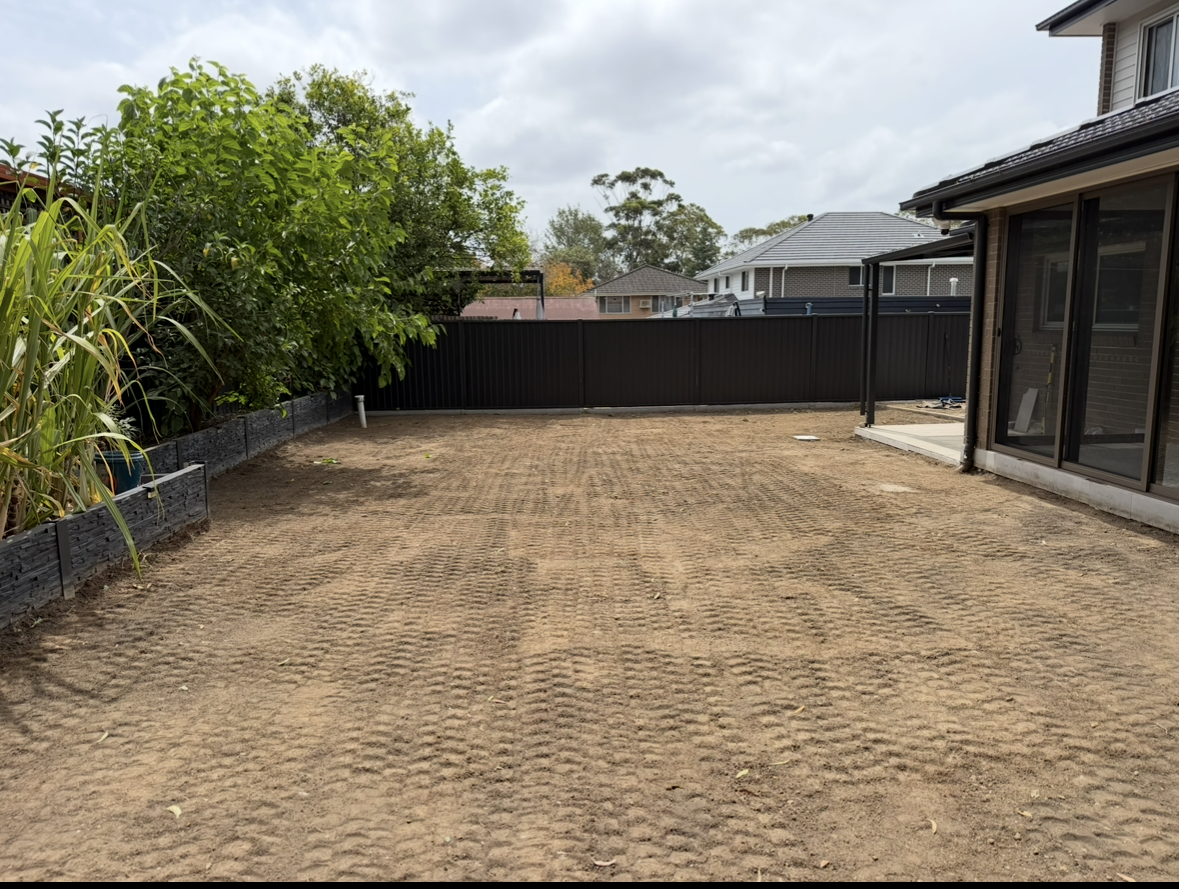 A backyard with freshly leveled dirt, side garden bed with plants on the left, and a black metal fence at the back. There is a house on the right with a patio area.