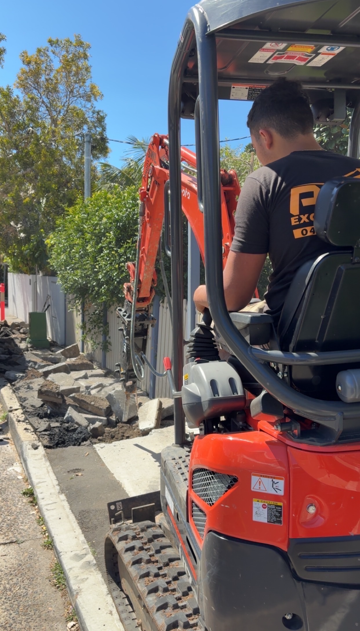 Construction worker operating a mini excavator on a sidewalk with broken concrete and rocks.