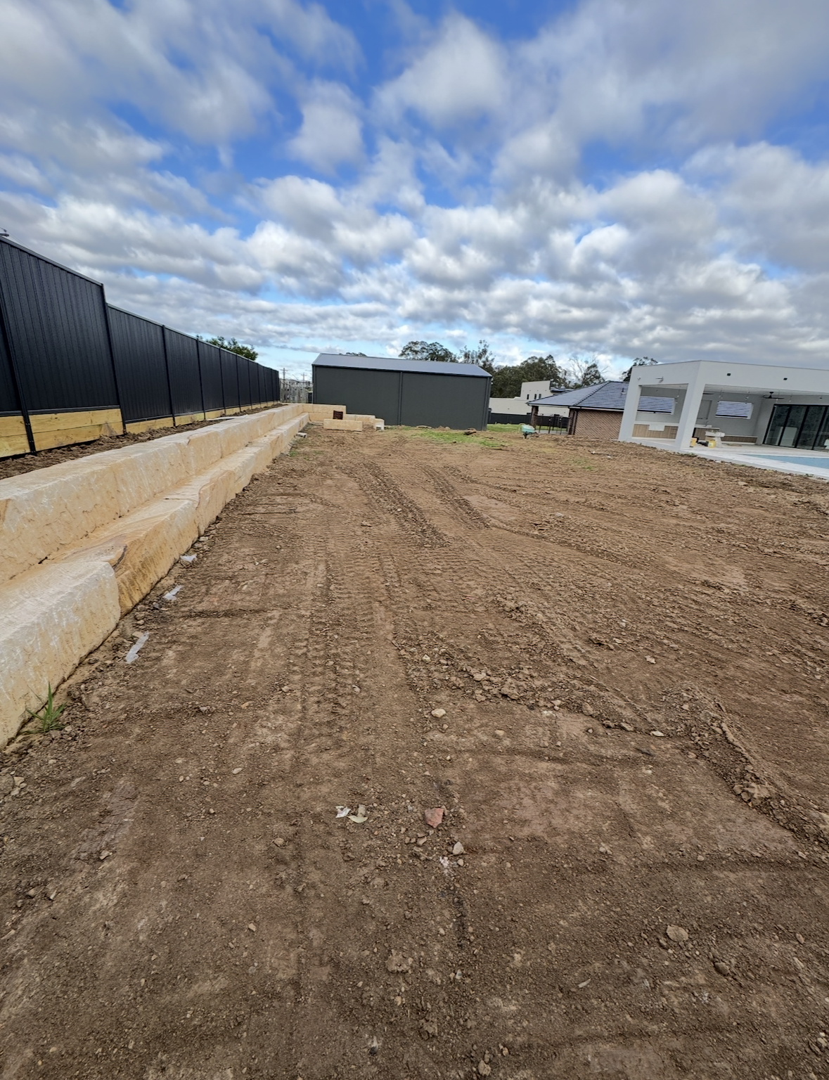 A cleared and leveled construction site with dirt ground, tire tracks, a black fence on the left, a small green shed in the background, and modern white houses on the right under a partly cloudy sky.