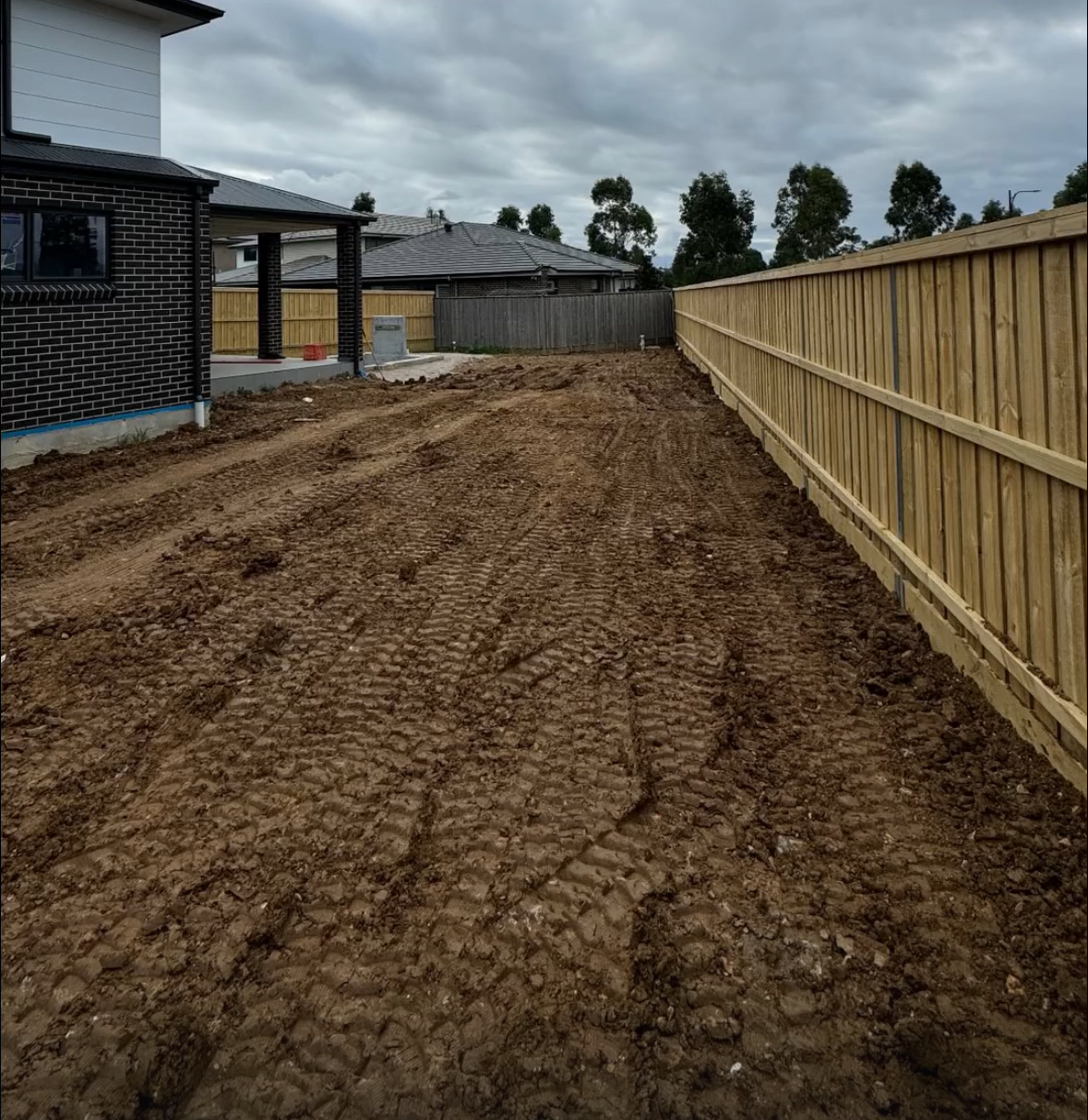 A dirt backyard with tire tracks, enclosed by a wooden fence on the right and houses in the background under cloudy sky.
