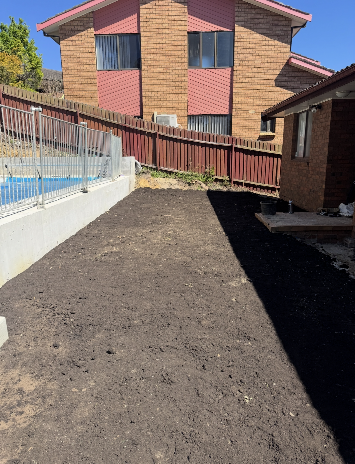A backyard with freshly leveled dark soil, bordered by a wooden fence and concrete edging, with a brick house on the right and a neighboring house in the background.