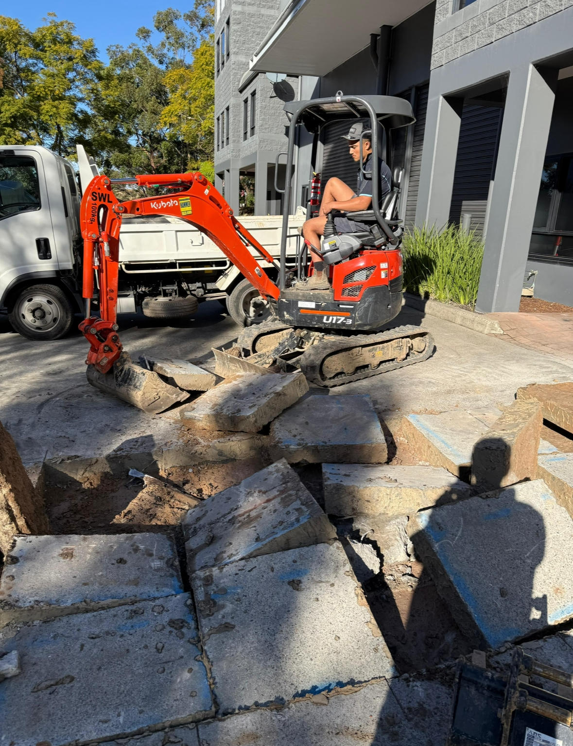 Construction worker operating a small red excavator on a city street, demolishing concrete pavement with broken pieces scattered around.