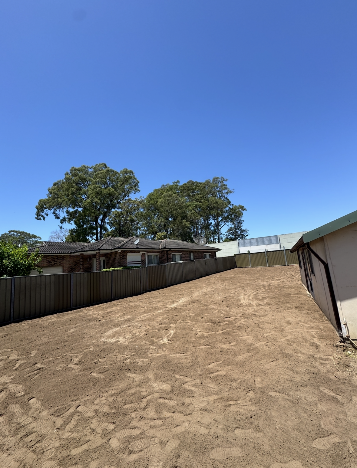 A backyard with a dirt ground, a metal fence, a brick house with satellite dishes, tall trees, and a clear blue sky.