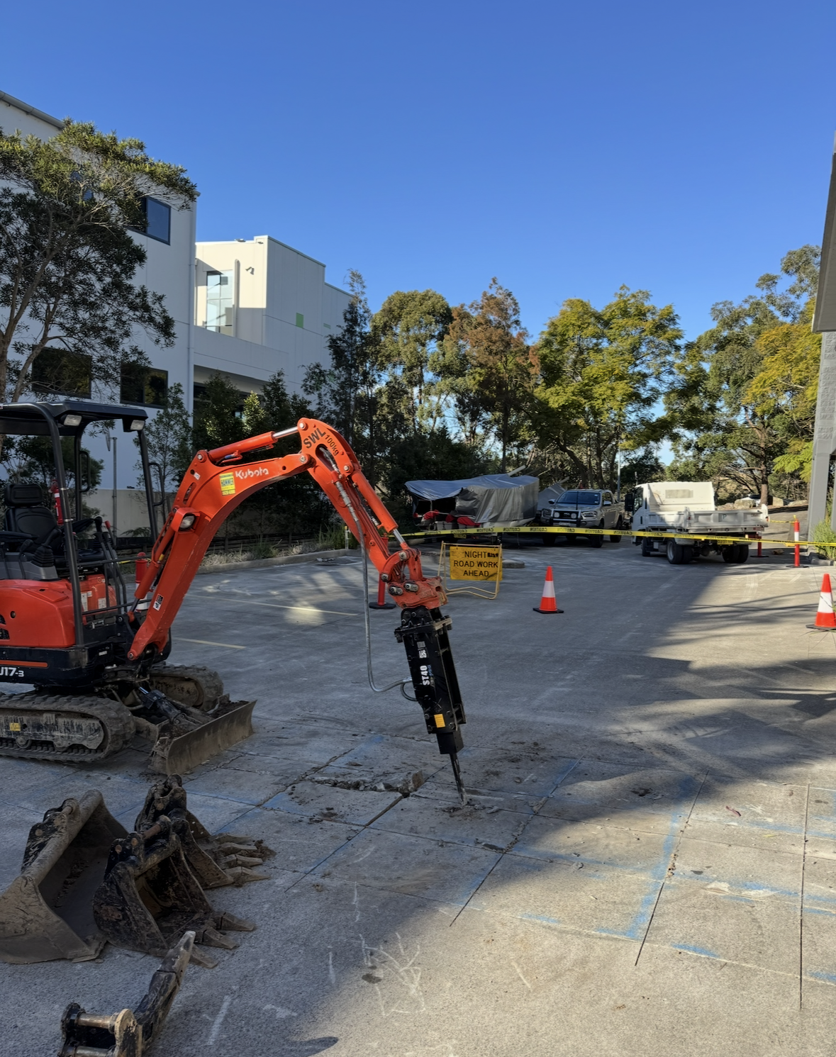 Construction site with a small excavator breaking pavement, orange traffic cones, and signs indicating night road work ahead. Residential buildings and parked vehicles are in the background.