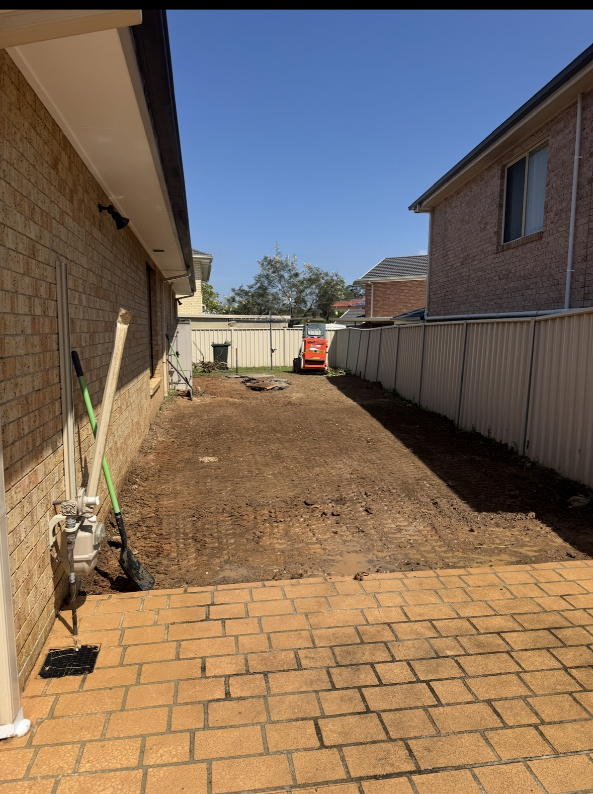 Backyard undergoing landscaping or yard renovation, with a paved brick patio in the foreground and bare earth extending to a fence and houses in the background, construction tools leaning against the house.