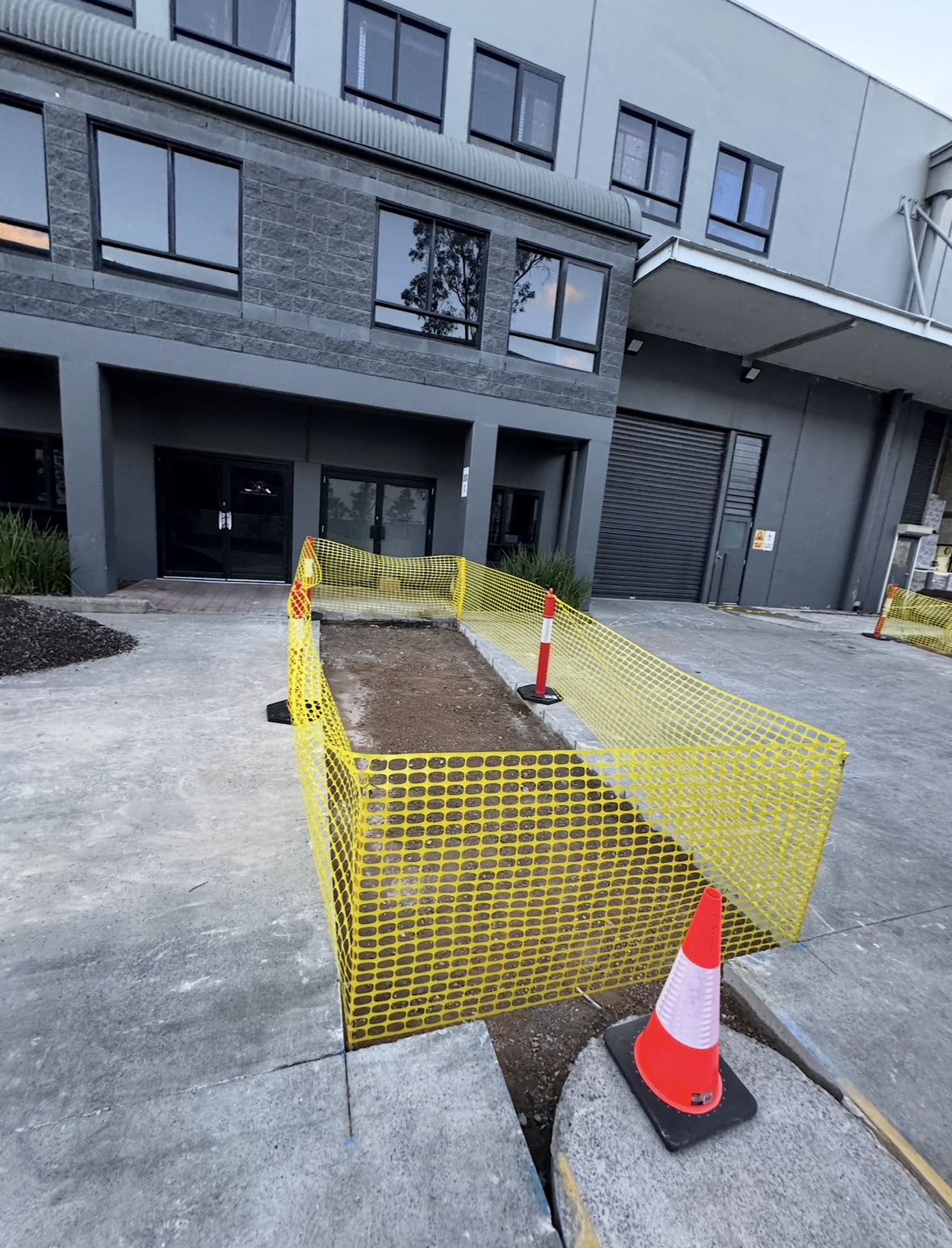 Construction site in front of a modern building with a fenced-off dirt area, orange and white traffic cone, and yellow safety barriers.