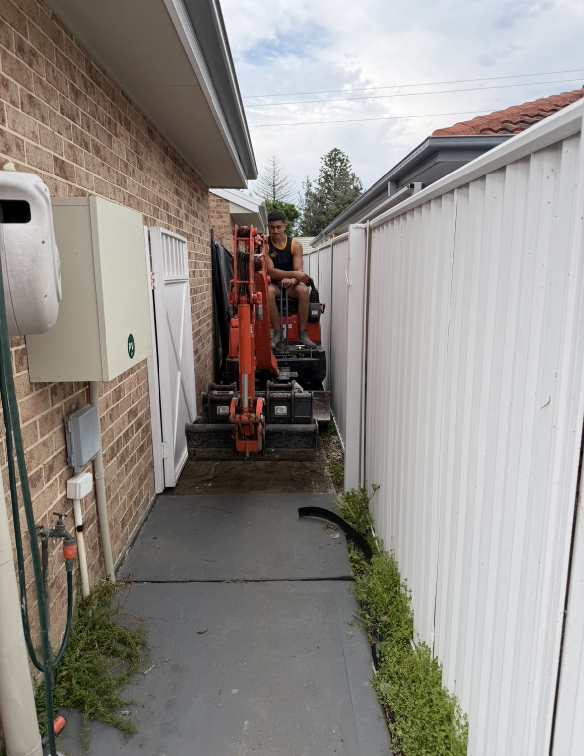 A person operating a small red excavator in a narrow outdoor space between a brick house and a white fence.
