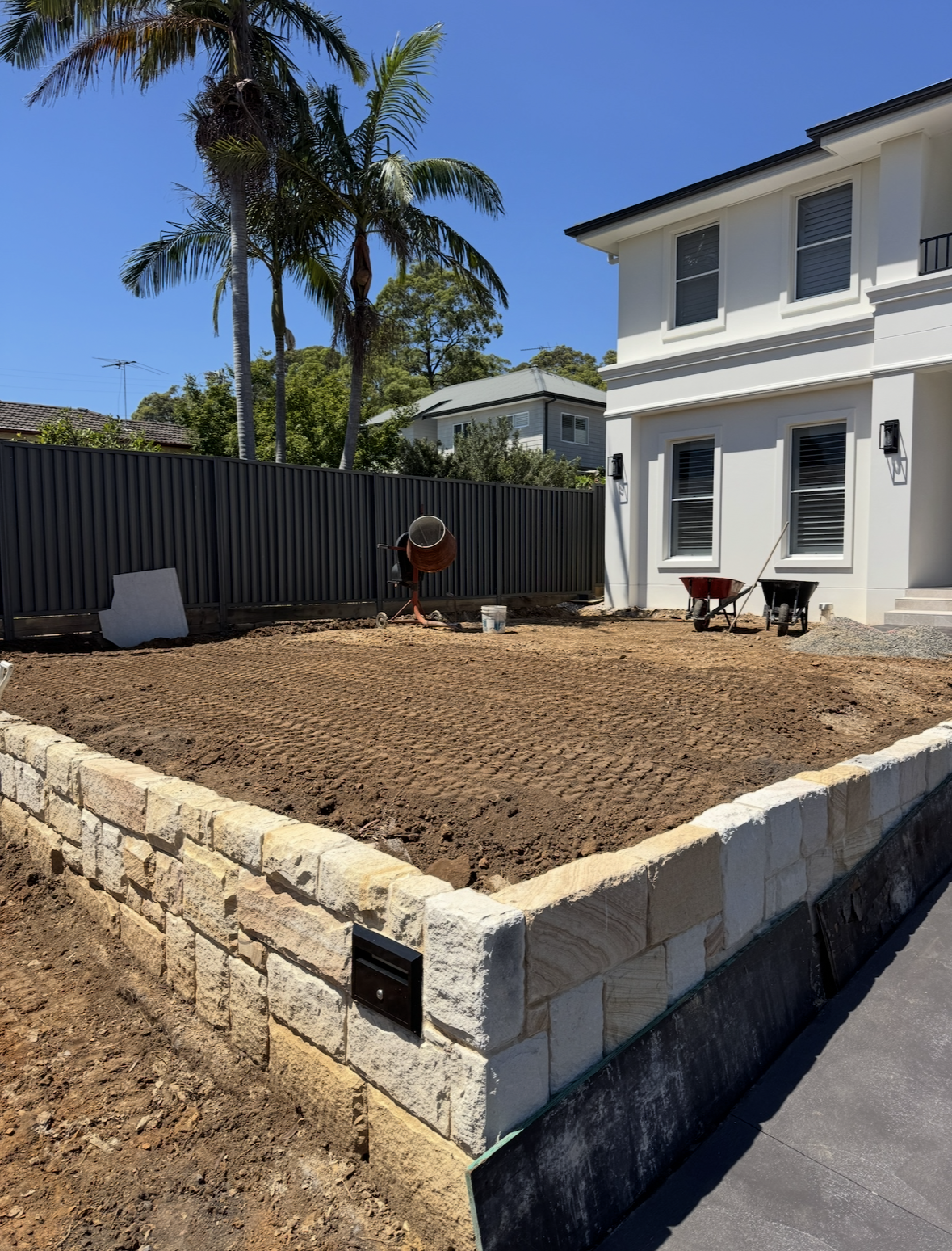 A backyard under construction with freshly tilled soil, a stone retaining wall, and a white two-story house with three windows facing the yard. Two wheelbarrows, a wheelbarrow full of gravel, and construction tools are present. Tall palm trees and ot