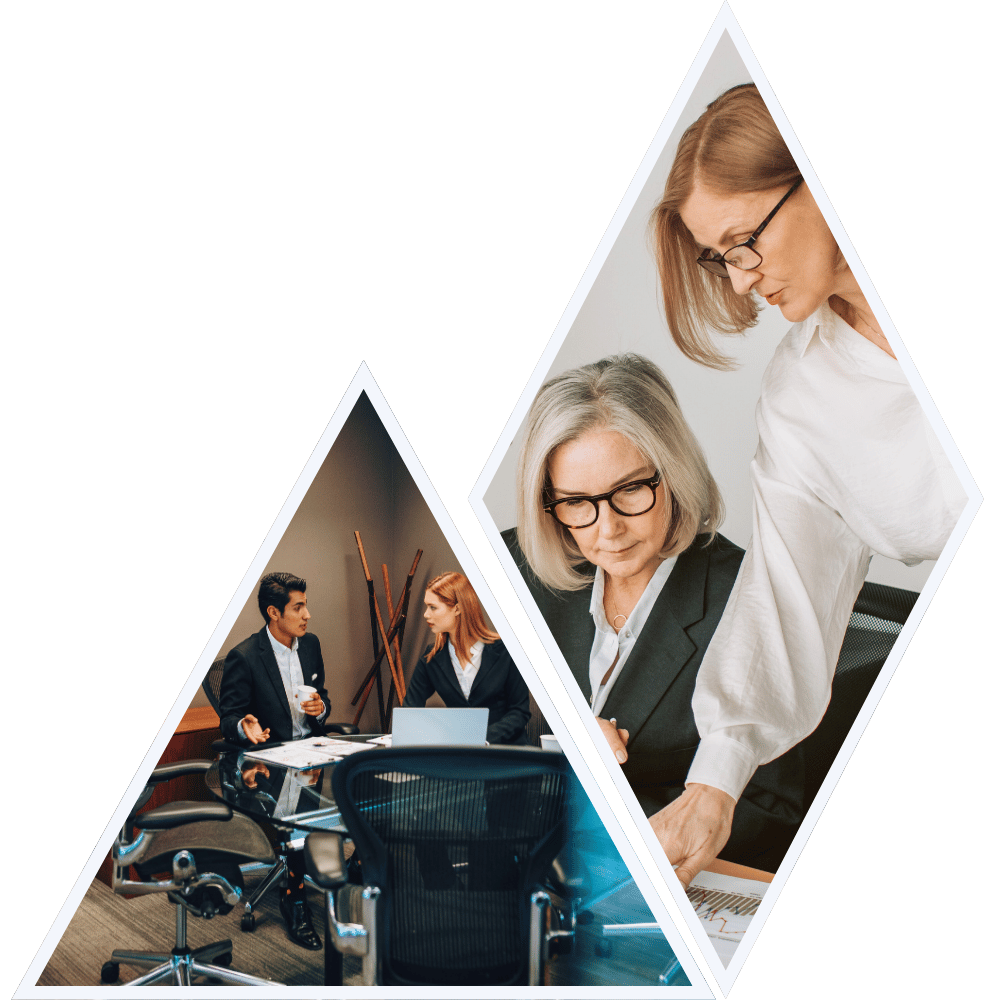 Two women in business attire reviewing documents at a meeting table, one is sitting and the other is standing, in a professional office setting.