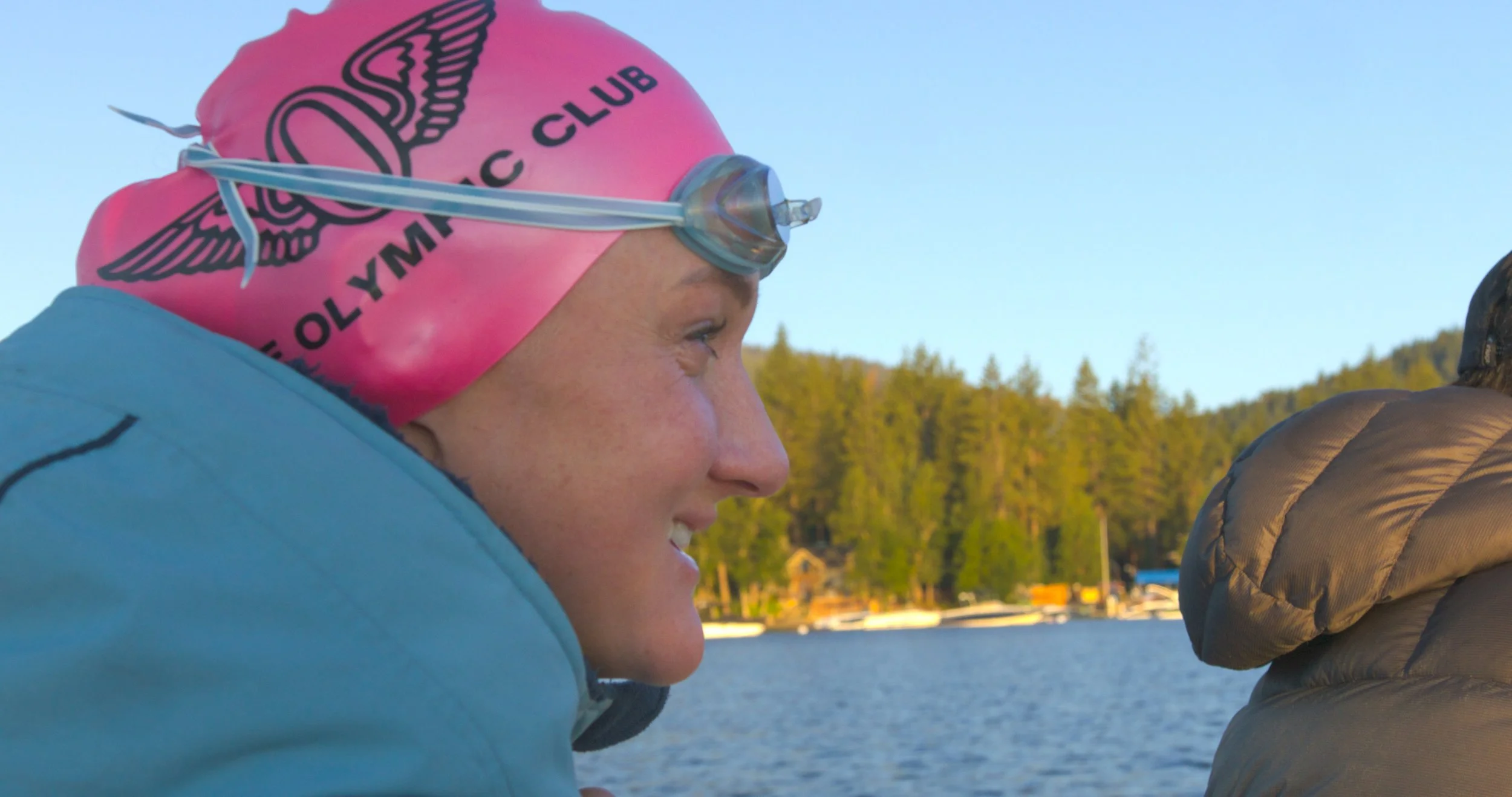 Close-up of a woman smiling wearing a pink swim cap with logo and goggles, outdoors near water and trees.