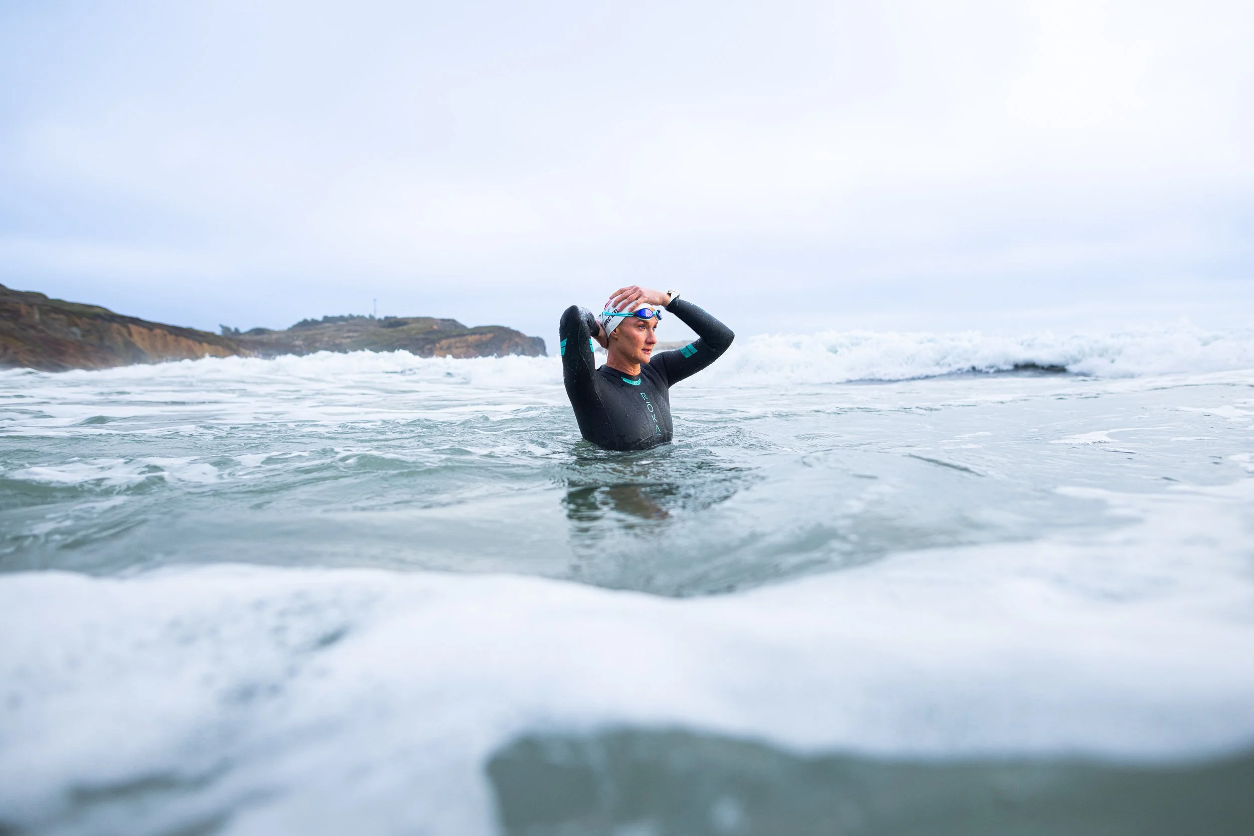 Woman in a wetsuit standing in the ocean, adjusting her goggles, with waves and rocky cliffs in the background.