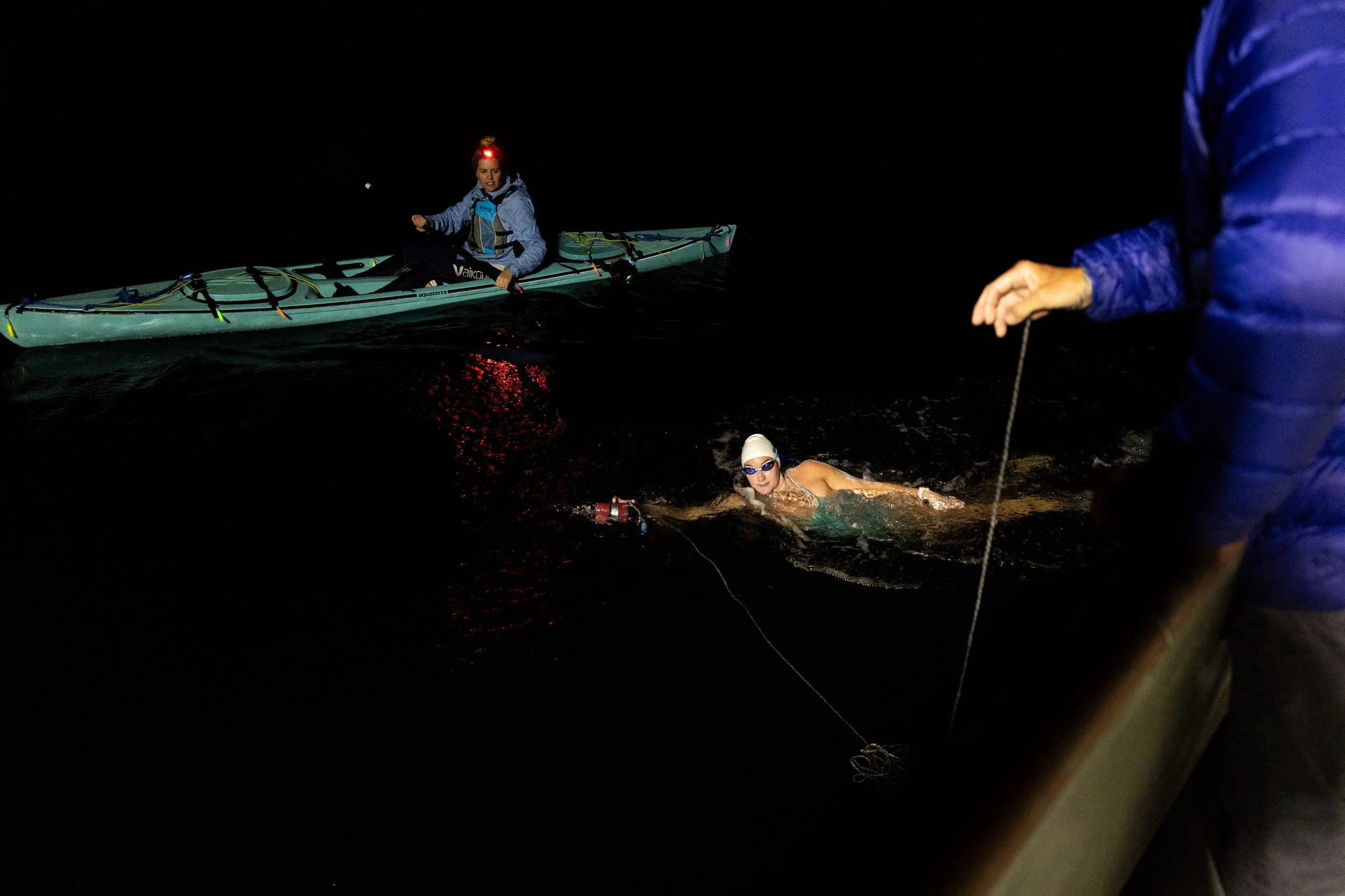 Nighttime image of a swimmer in a swim cap and goggles being assisted to a boat by a person holding a rope, with another person paddling a kayak in the background.