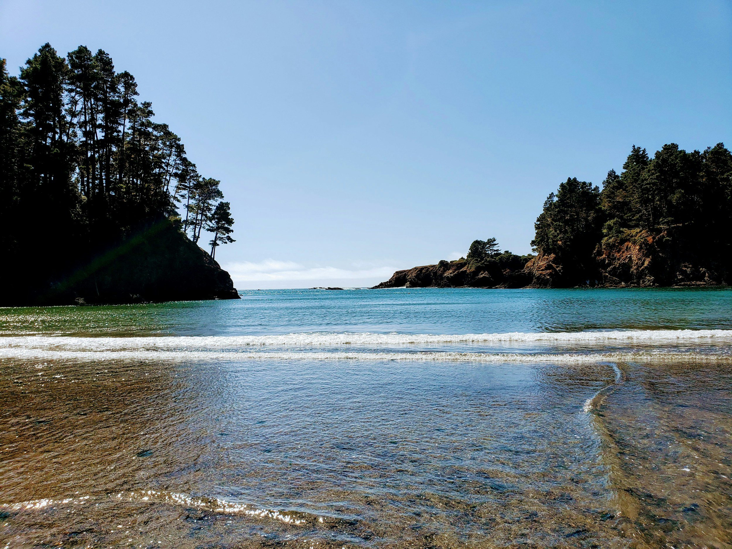 A scenic view of a beach with clear blue water, rocky cliffs covered with trees on both sides, and a bright blue sky overhead.