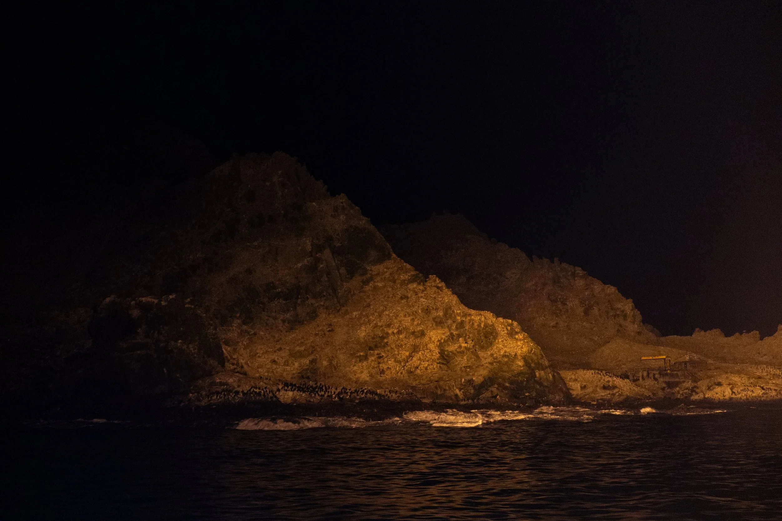 Nighttime view of rocky coastline with mountains, illuminated by warm artificial light, with water in the foreground.
