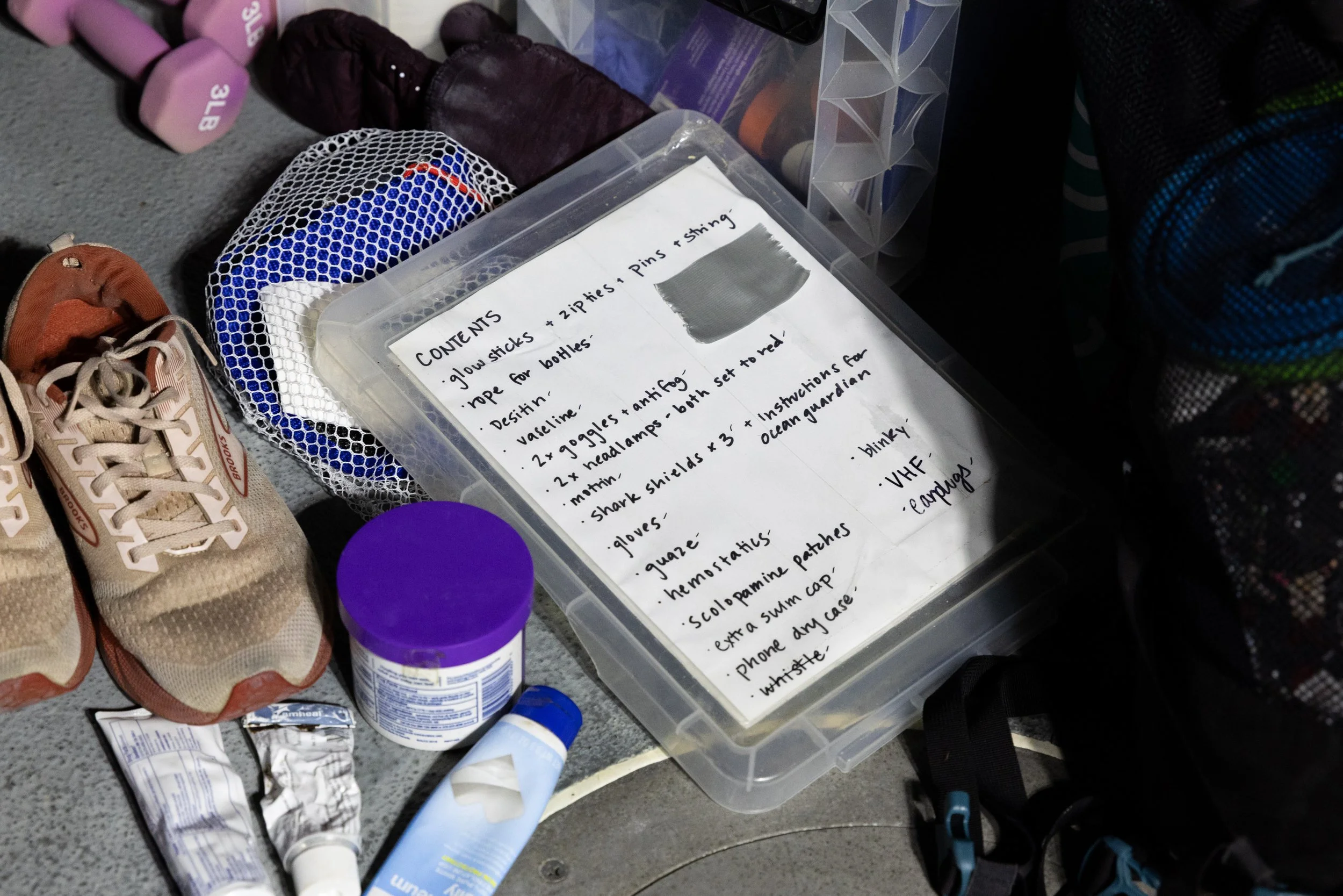 A clear plastic container with a handwritten list of contents for a first aid kit on top, including items such as glow sticks, Vaseline, goggles, headlamps, shark shields, gloves, gauze, hemastat, extras for a swim cap, phone dry case, whistle, a gra