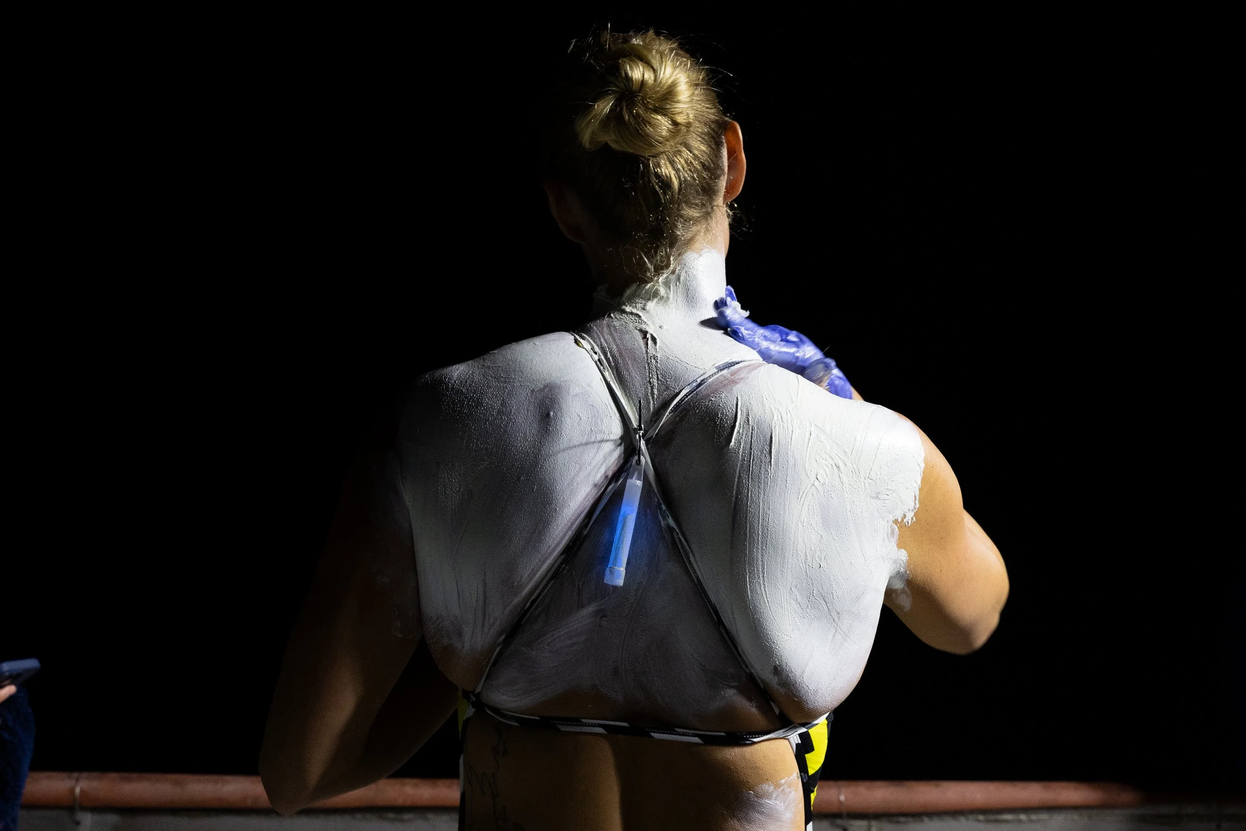 A woman with blonde hair in a bun is seen from behind, exposing her back. She is covered in white chalk or clay and is wearing a harness. She has blue gloves on one hand and appears to be preparing for or in the middle of a physical activity, possibl