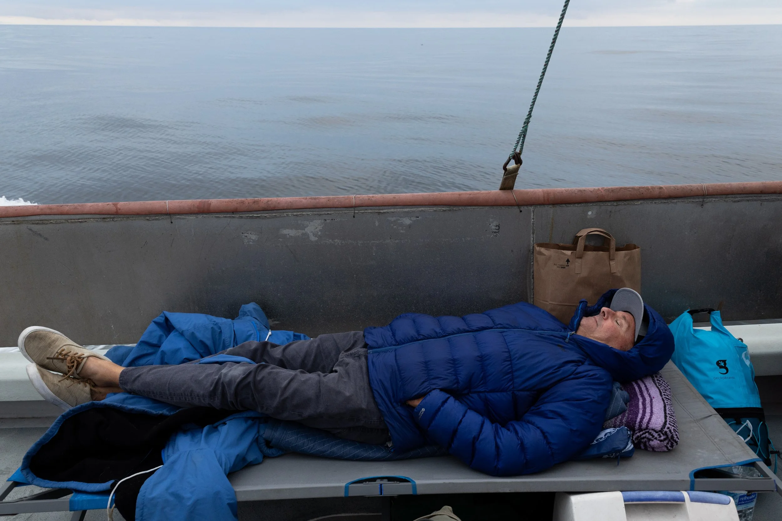 A man sleeping on a camping cot on a boat with water in the background, wearing a blue jacket and cap, with a pillow and bags nearby.