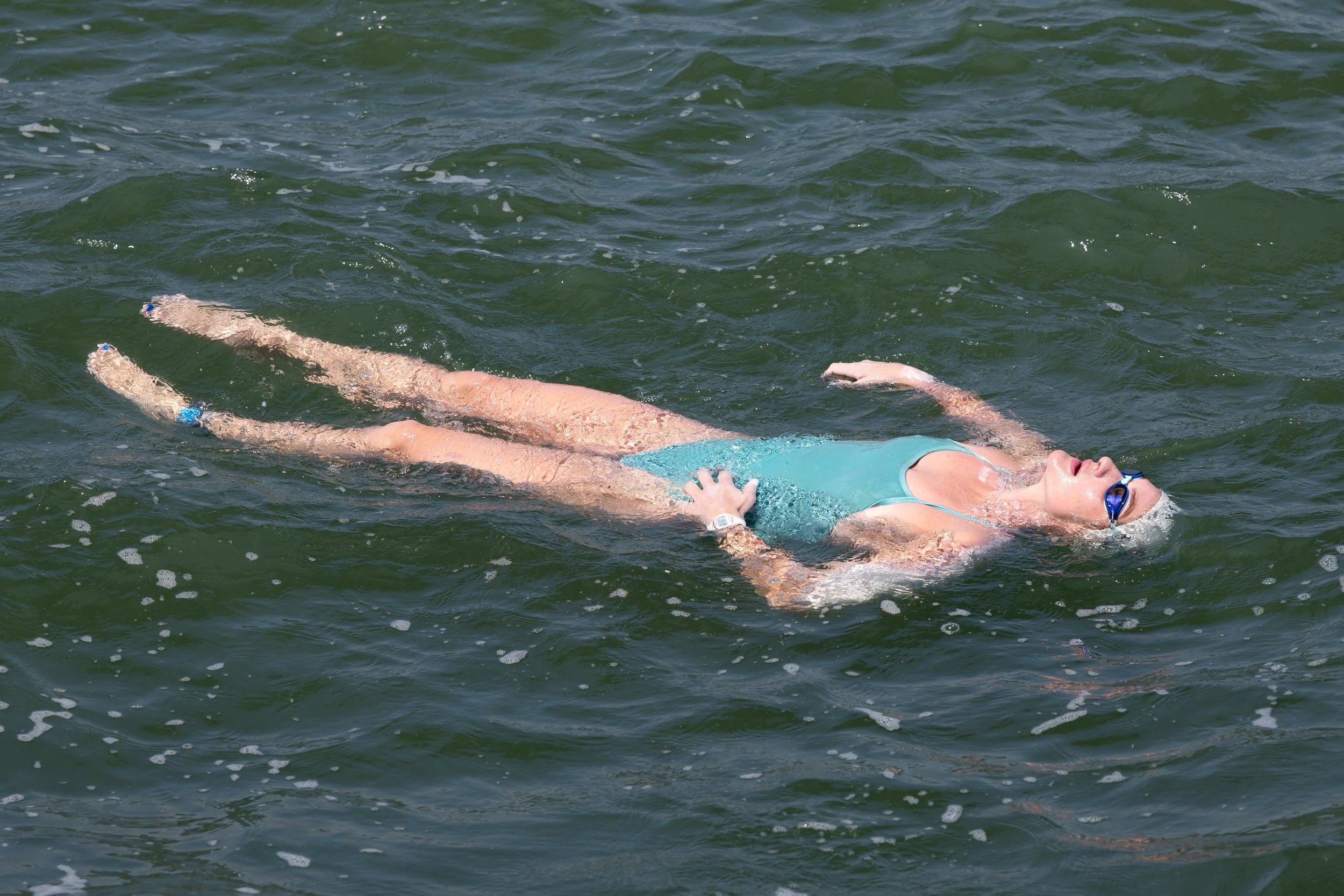 A woman swimming in a body of water, wearing a teal swimsuit and blue goggles, floating on her back.