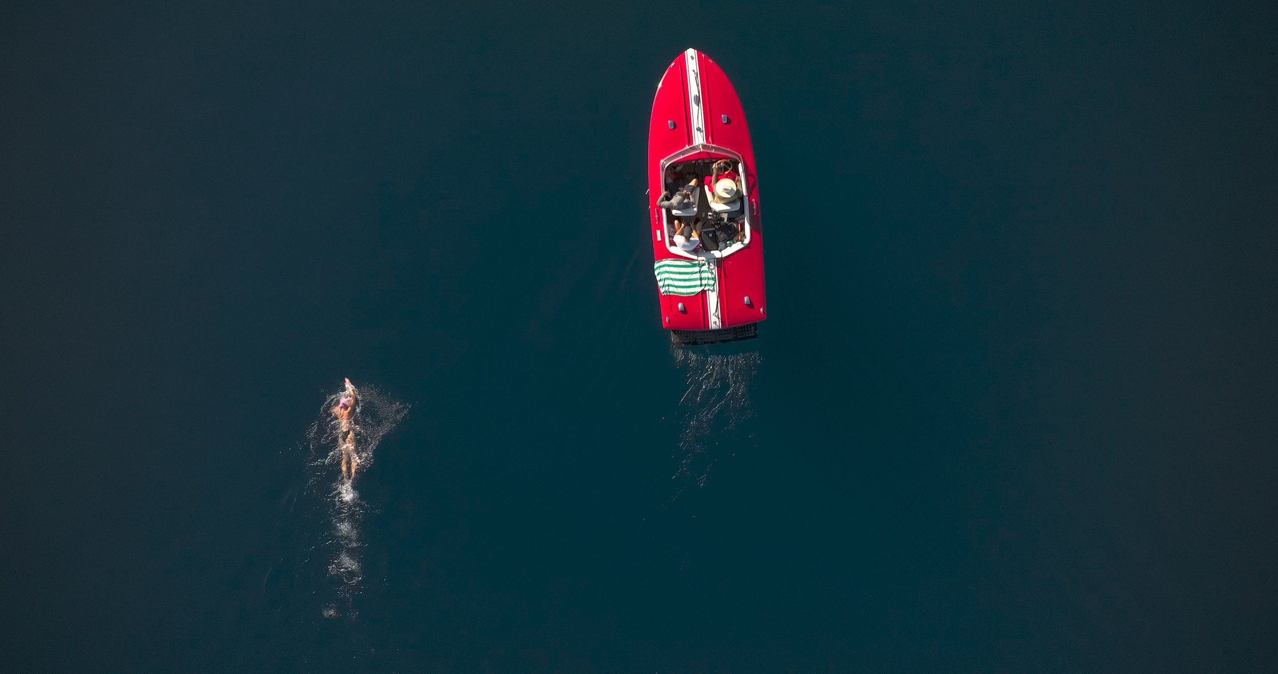An aerial view of a red boat with two people on board, and a swimmer in open water nearby.
