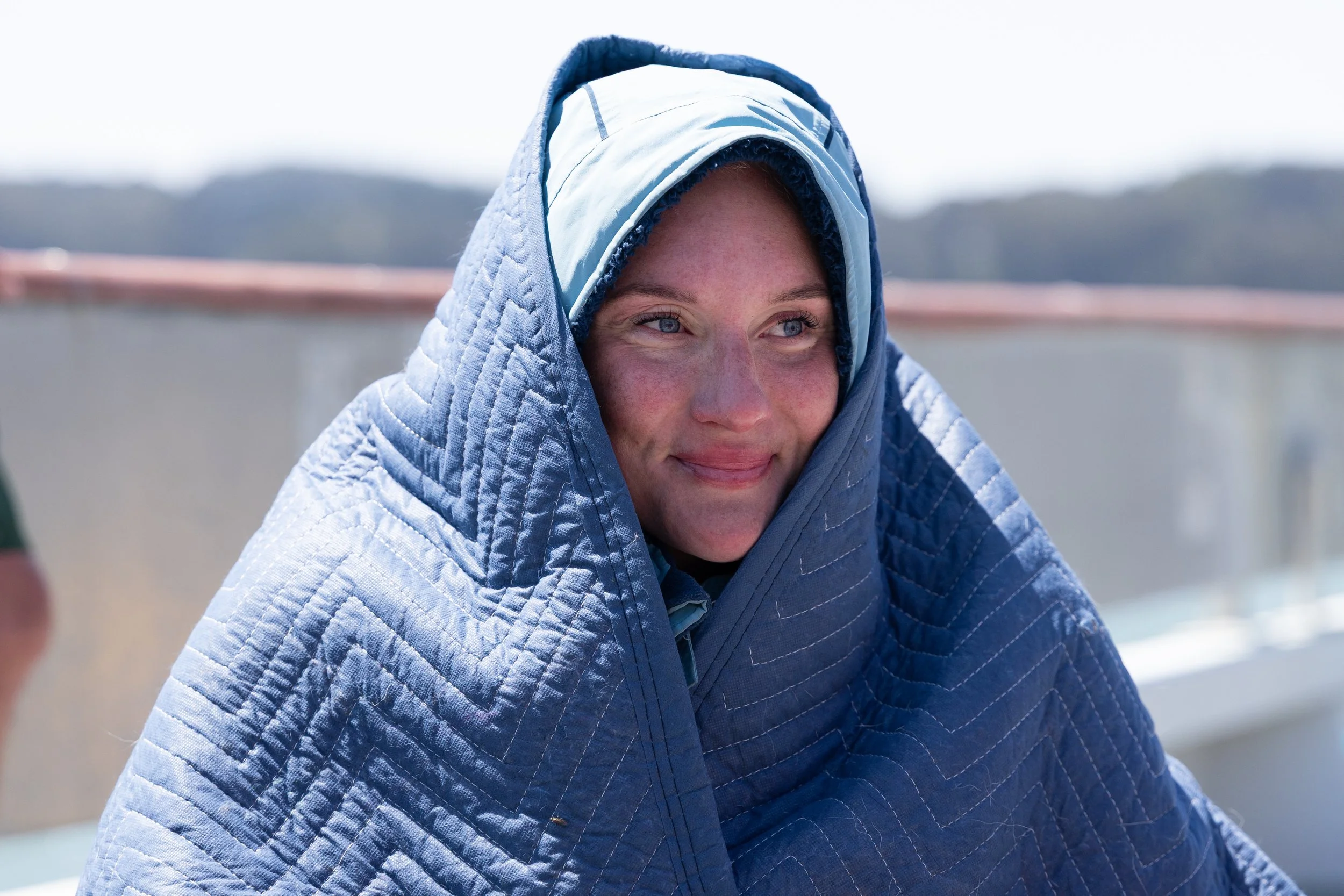 A woman with blue eyes, smiling, wrapped warmly in a blue quilted blanket on a cold or windy day outdoors.