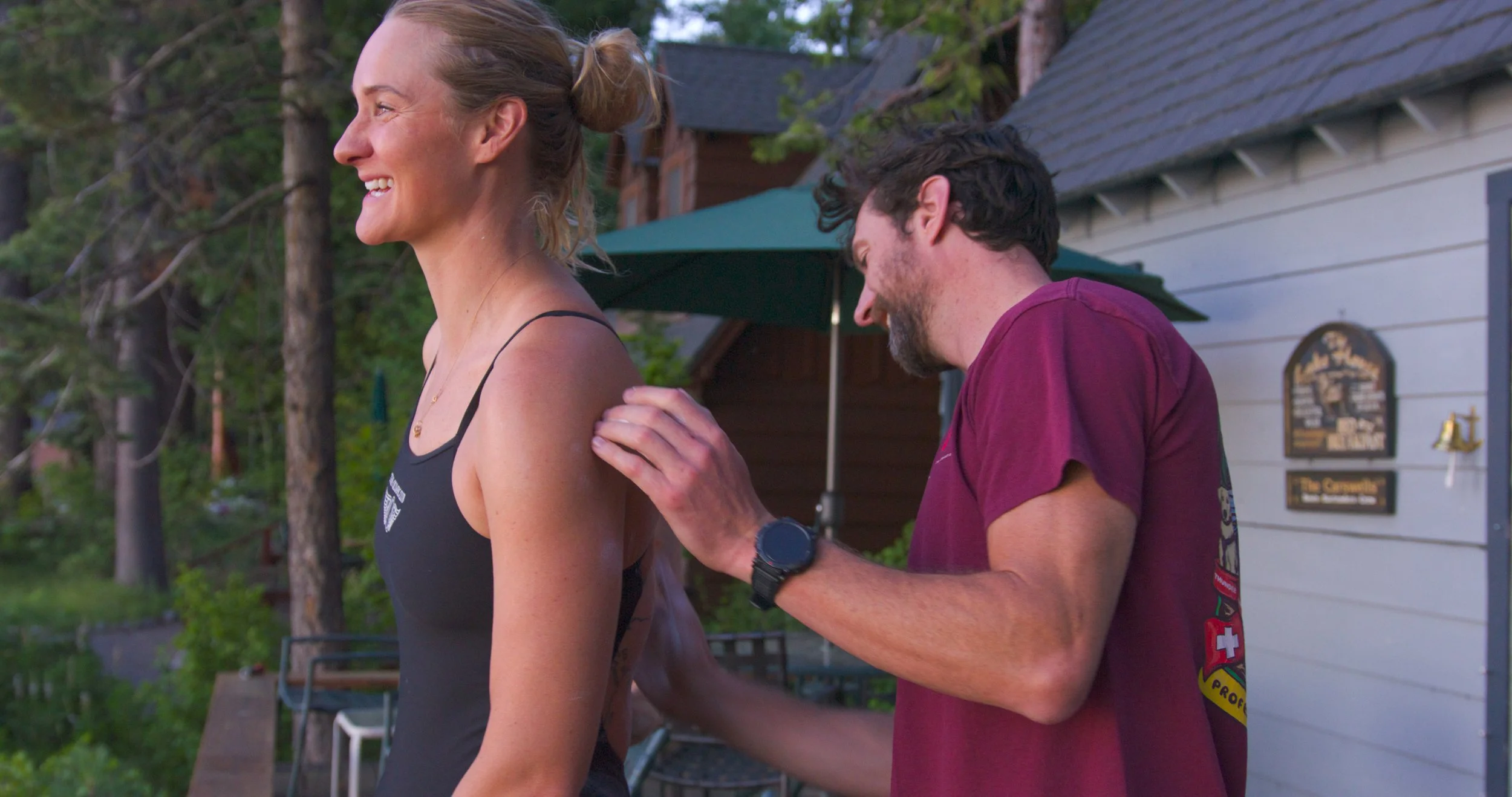 A woman with blonde hair in a bun smiling, wearing a black sports top and a necklace, is standing outdoors next to a man with dark hair and beard, wearing a maroon t-shirt, who appears to be adjusting her top. They are in front of a white house with 