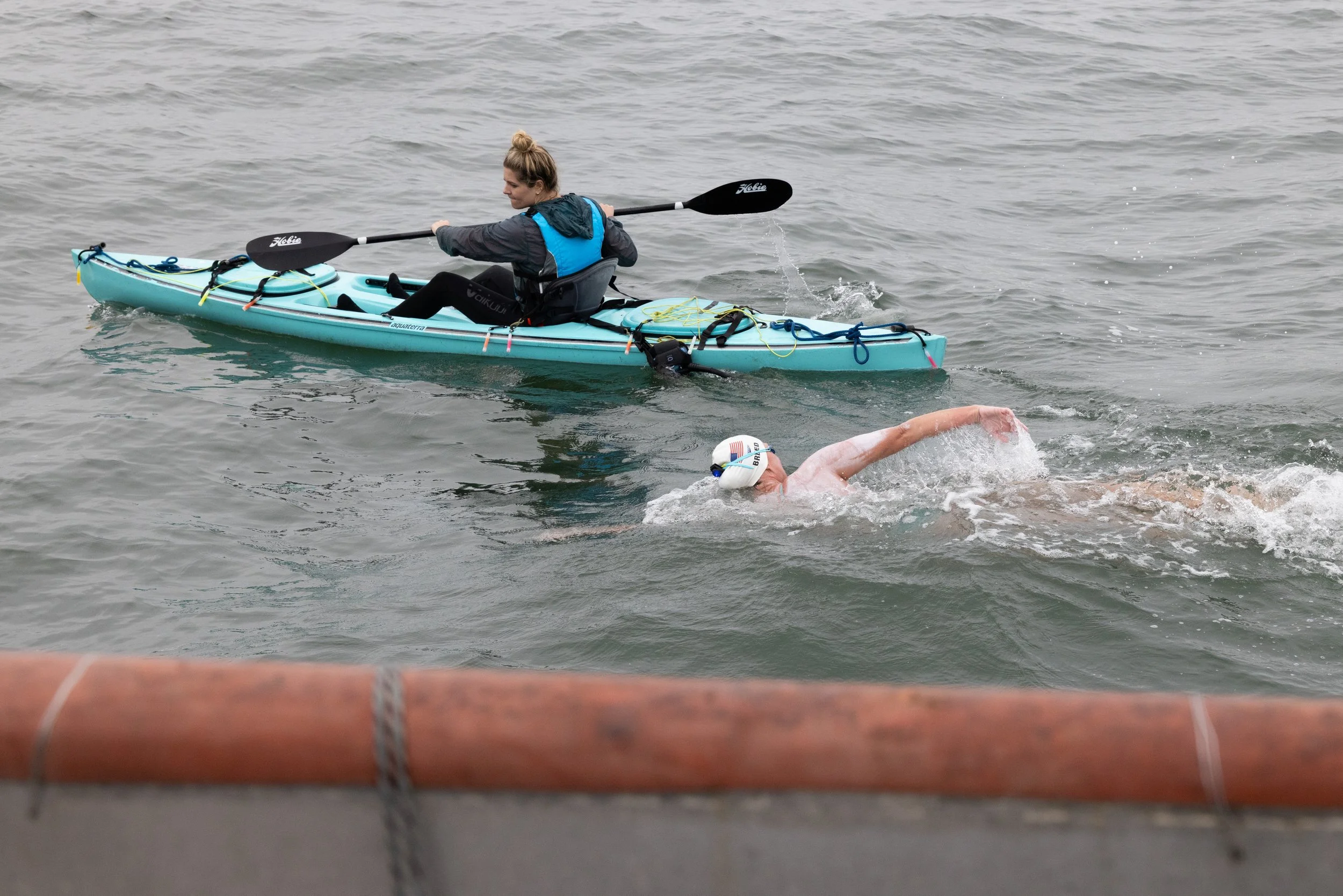 A person in a kayak with a double-bladed paddle on the water, and a swimmer in a swim cap swimming nearby.