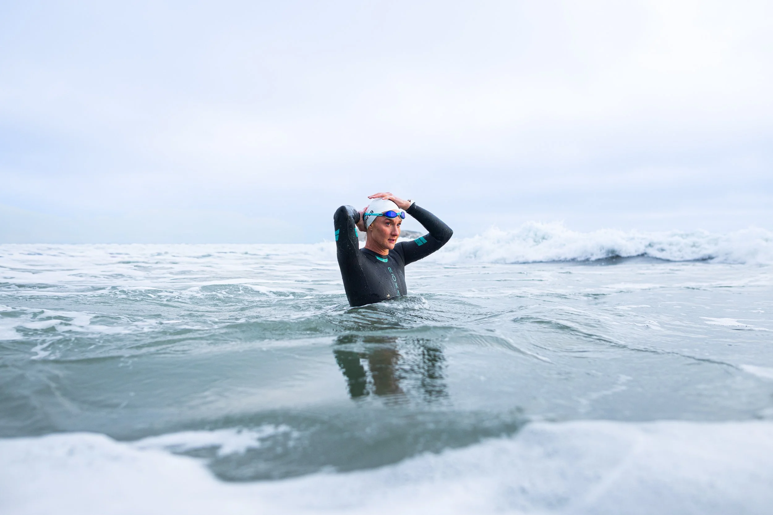 A woman in wetsuit and swim cap stands in the ocean, adjusting her swim cap after swimming.