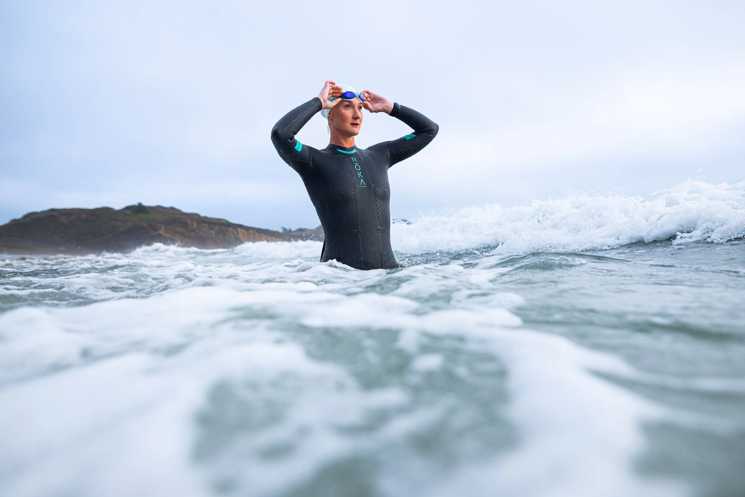 Woman in a wetsuit putting on goggles while standing in the ocean with a rocky island on the horizon.