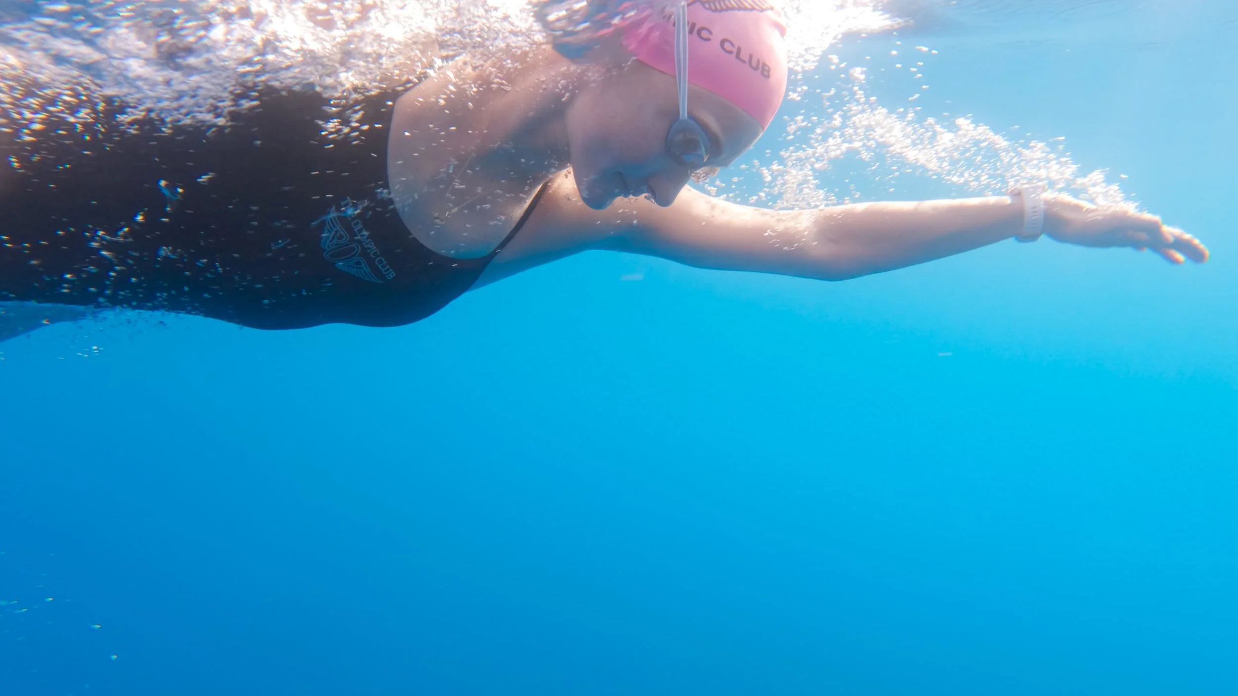 Female swimmer wearing a pink swim cap and goggles swims underwater in a blue swimming pool, with her arm extended forward.
