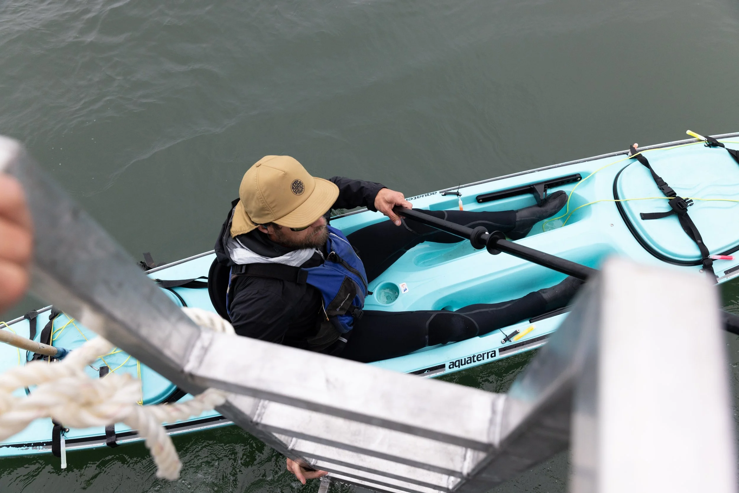 A man in a beige cap and black jacket is sitting in a blue kayak, holding a paddle, as he prepares to climb onto a boat via a metal ladder over water.