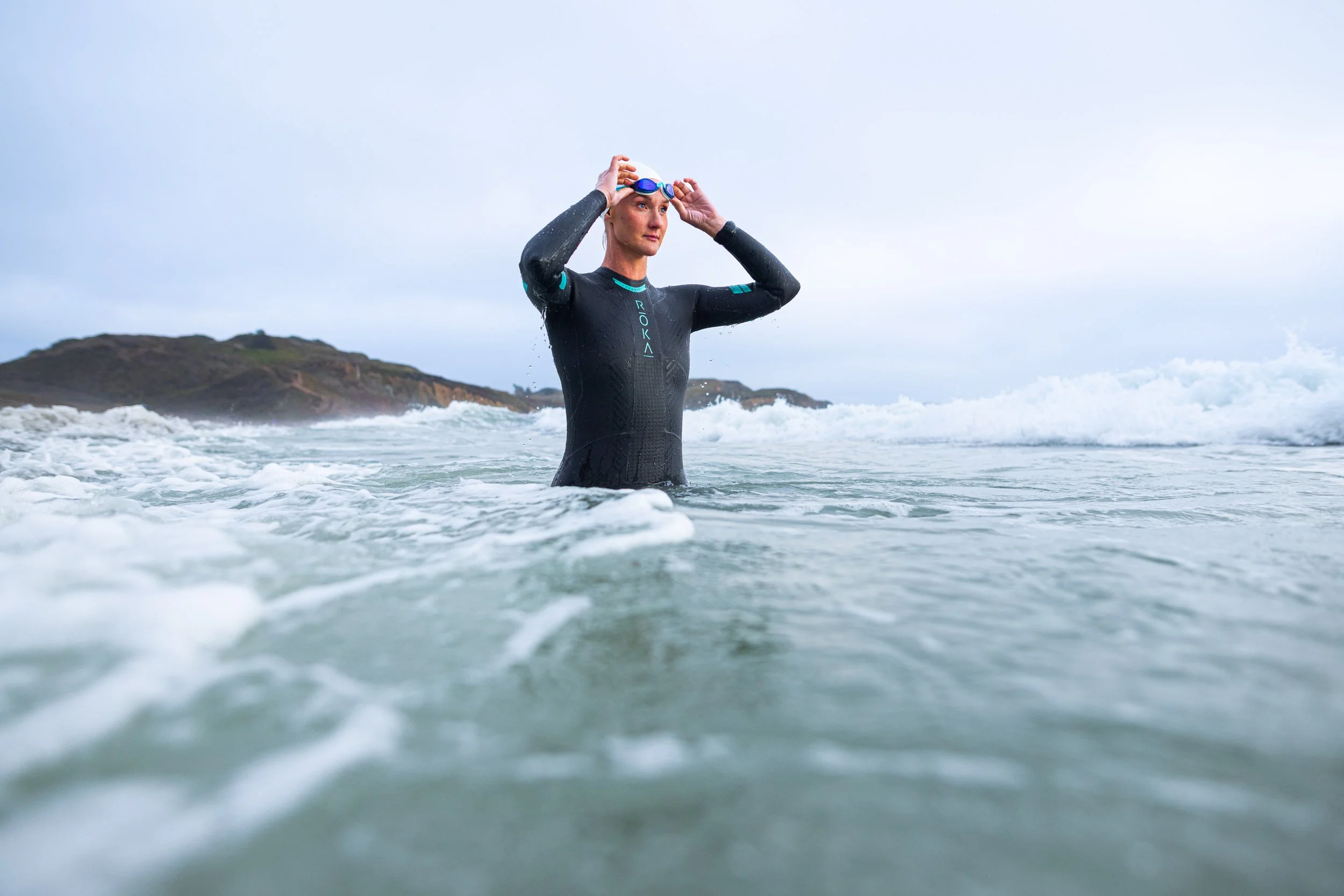 Woman in wetsuit adjusting goggles in ocean with hills in background.