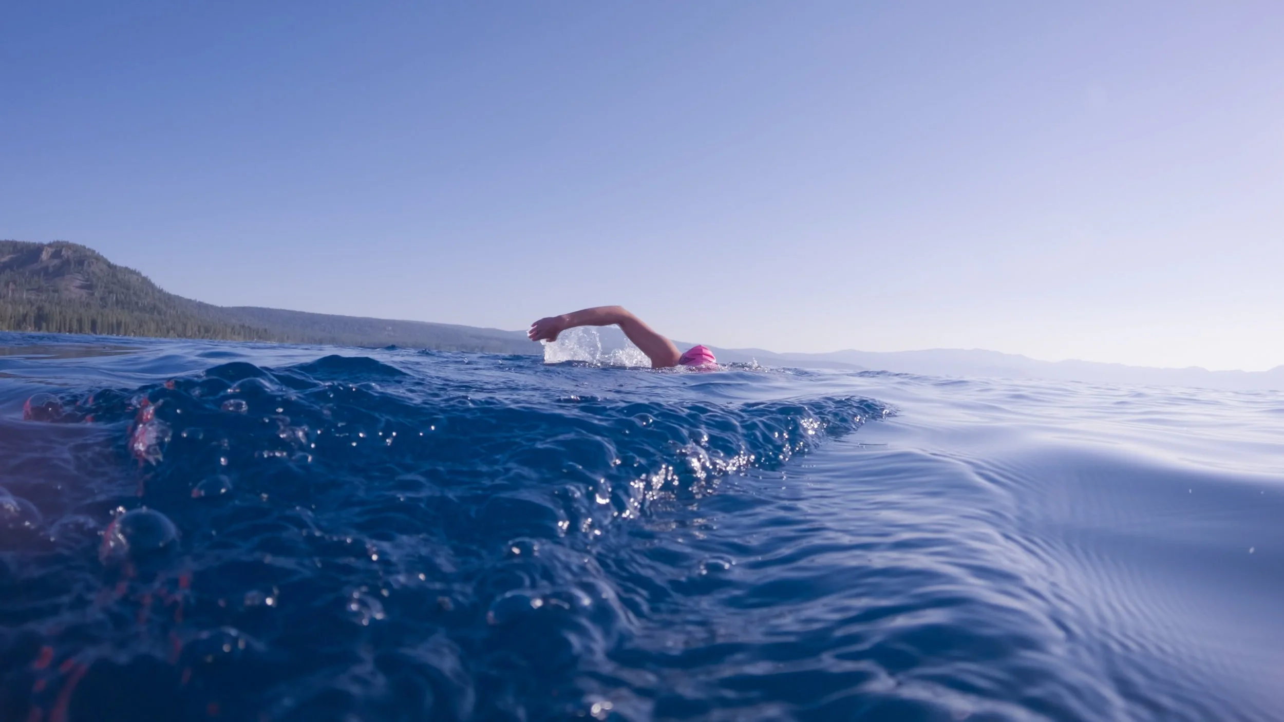 Swimmer in a pink cap swimming in open water under a clear blue sky with distant mountains on the horizon.