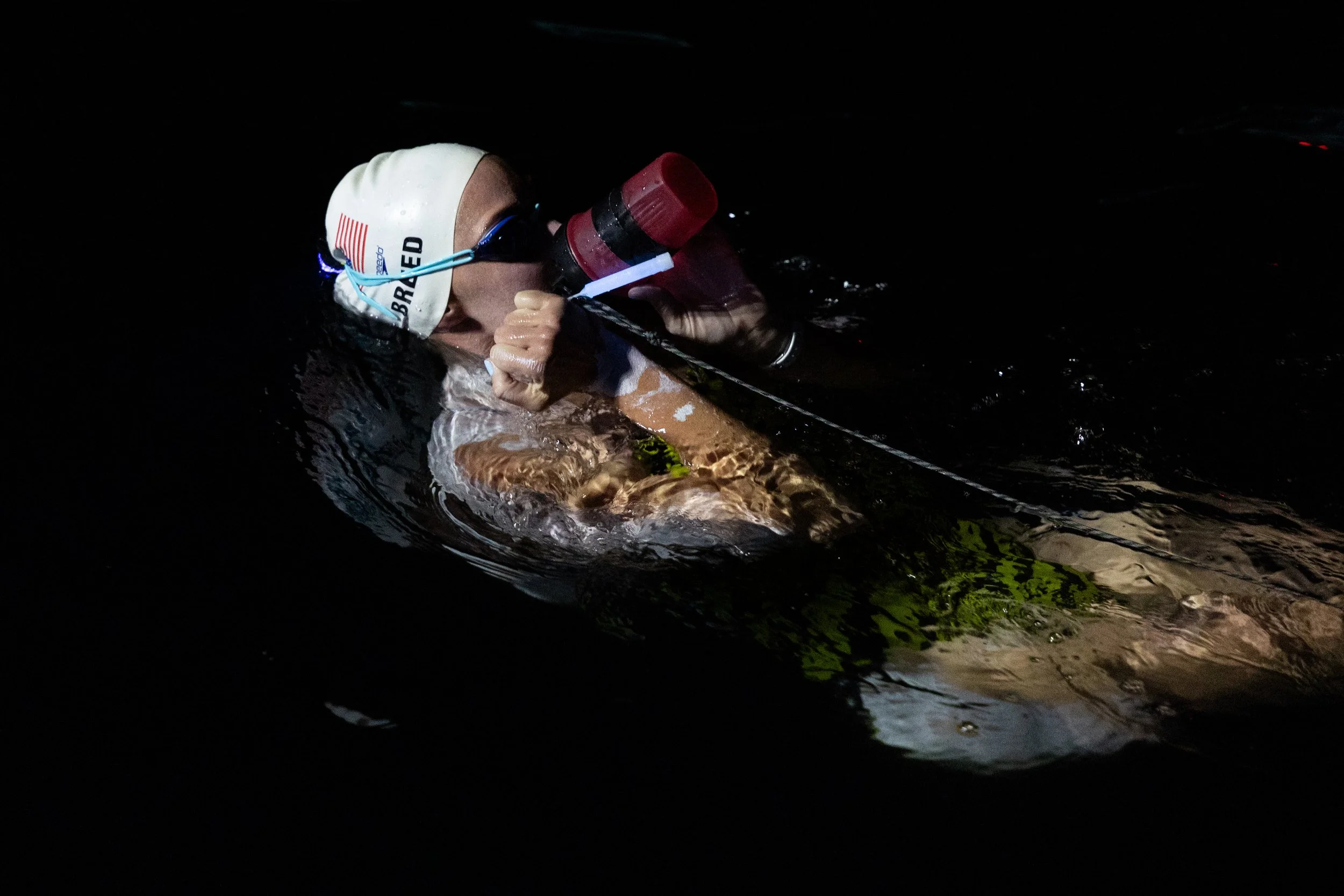 A swimmer wearing a white swim cap and dark goggles, holding a red water bottle, swimming in dark water at night.