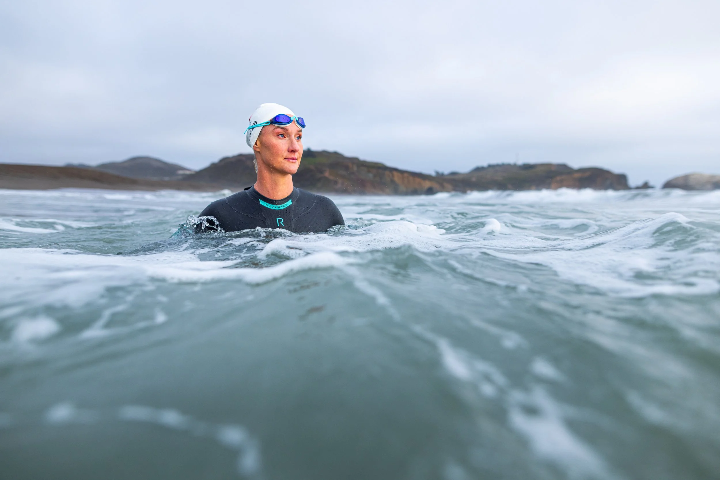 A woman in a wetsuit and swimming cap with goggles stands in the ocean amidst waves, with hills and cloudy sky in the background.
