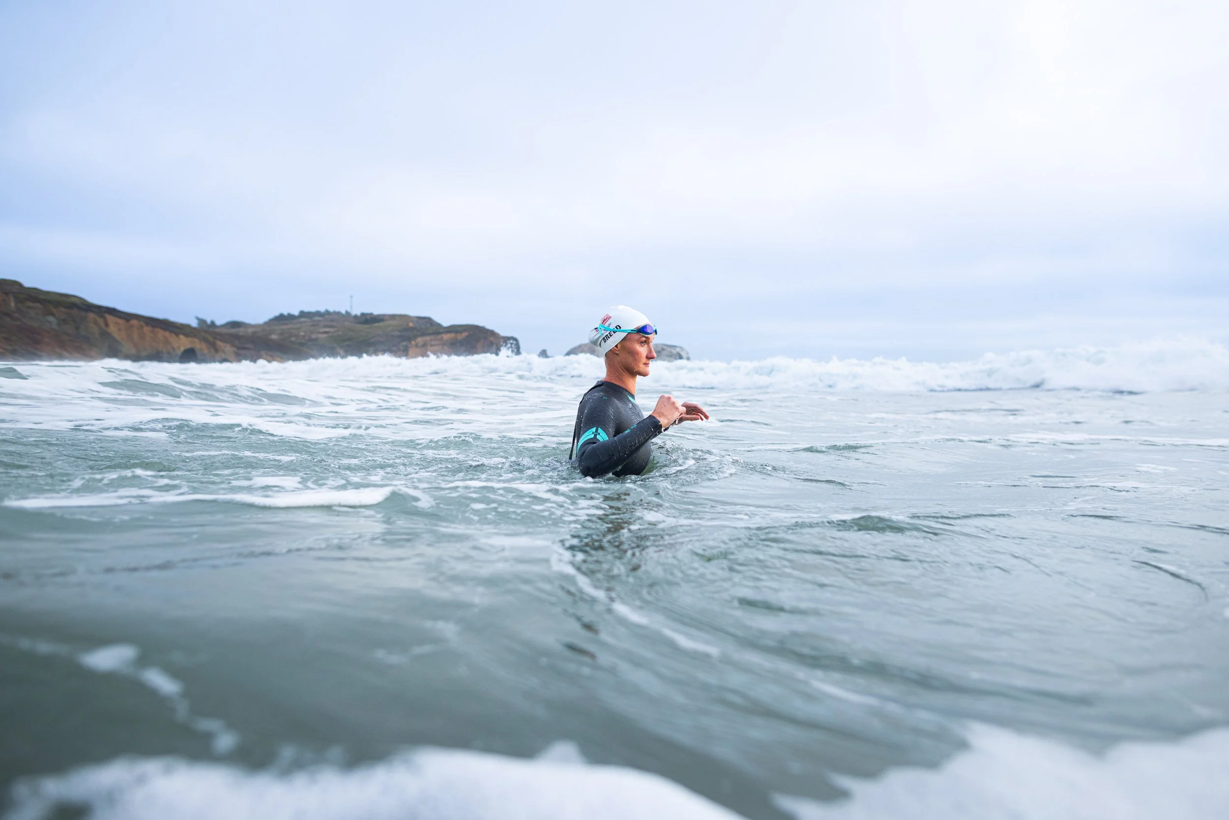 A swimmer in a wetsuit and swim cap standing in the ocean with a shoreline and cliffs in the background.