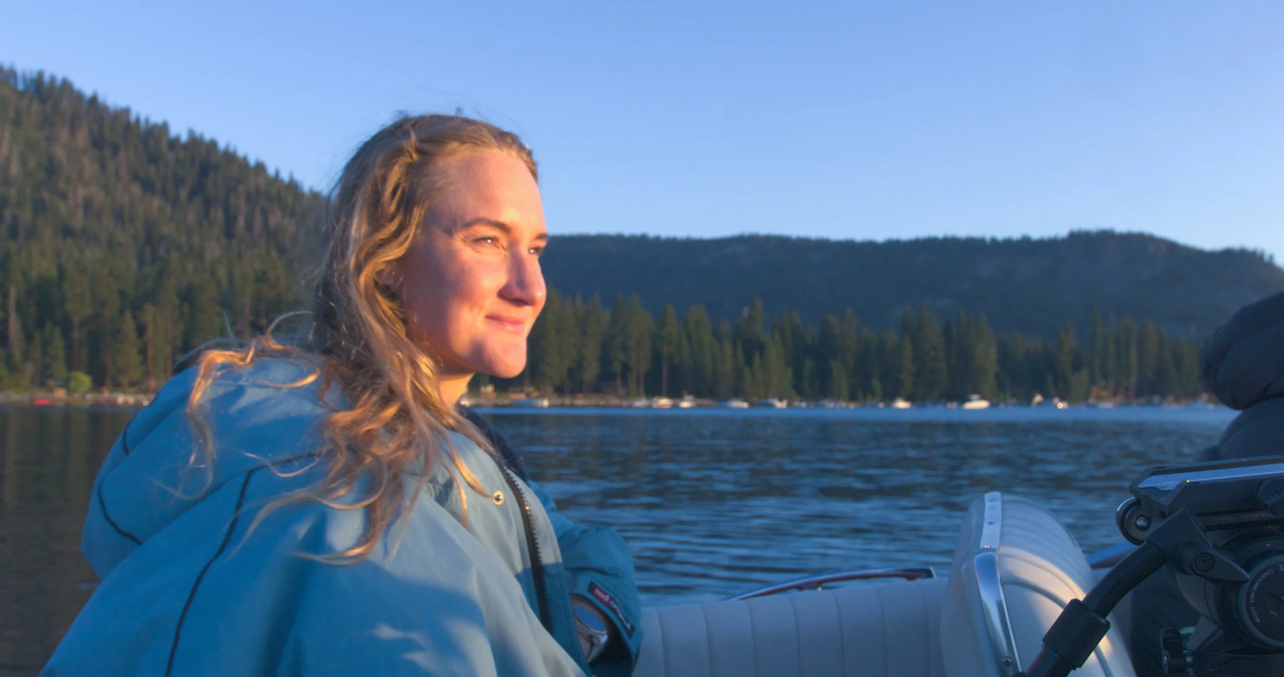 A woman with long curly blonde hair on a boat, smiling at sunset on a lake with a forested shoreline and mountains in the background.