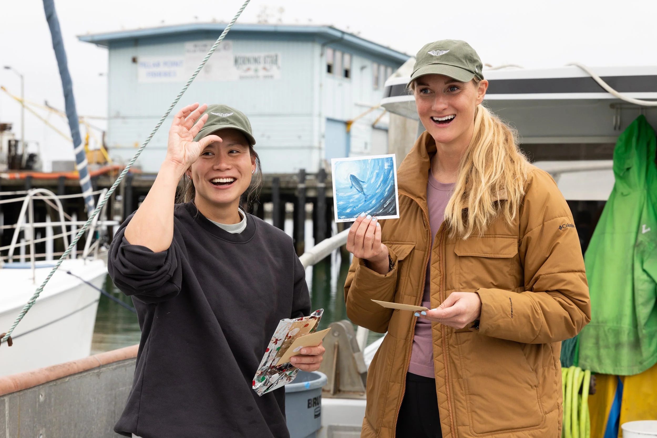 Two women smiling at a marina, one holding a photograph of a whale and the other holding a card, both wearing casual outdoor jackets and caps.