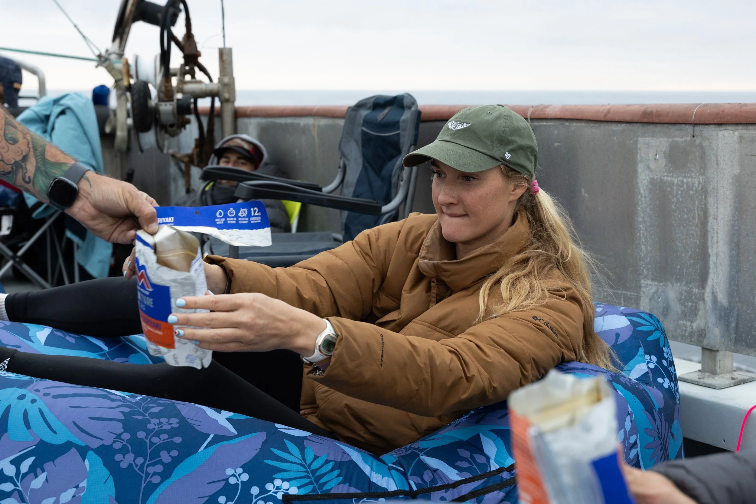A woman with blonde hair in a ponytail wearing a green cap and brown jacket sitting on a boat, receiving a packaged snack from someone. The boat has blue patterned seats, and there are other people in the background.