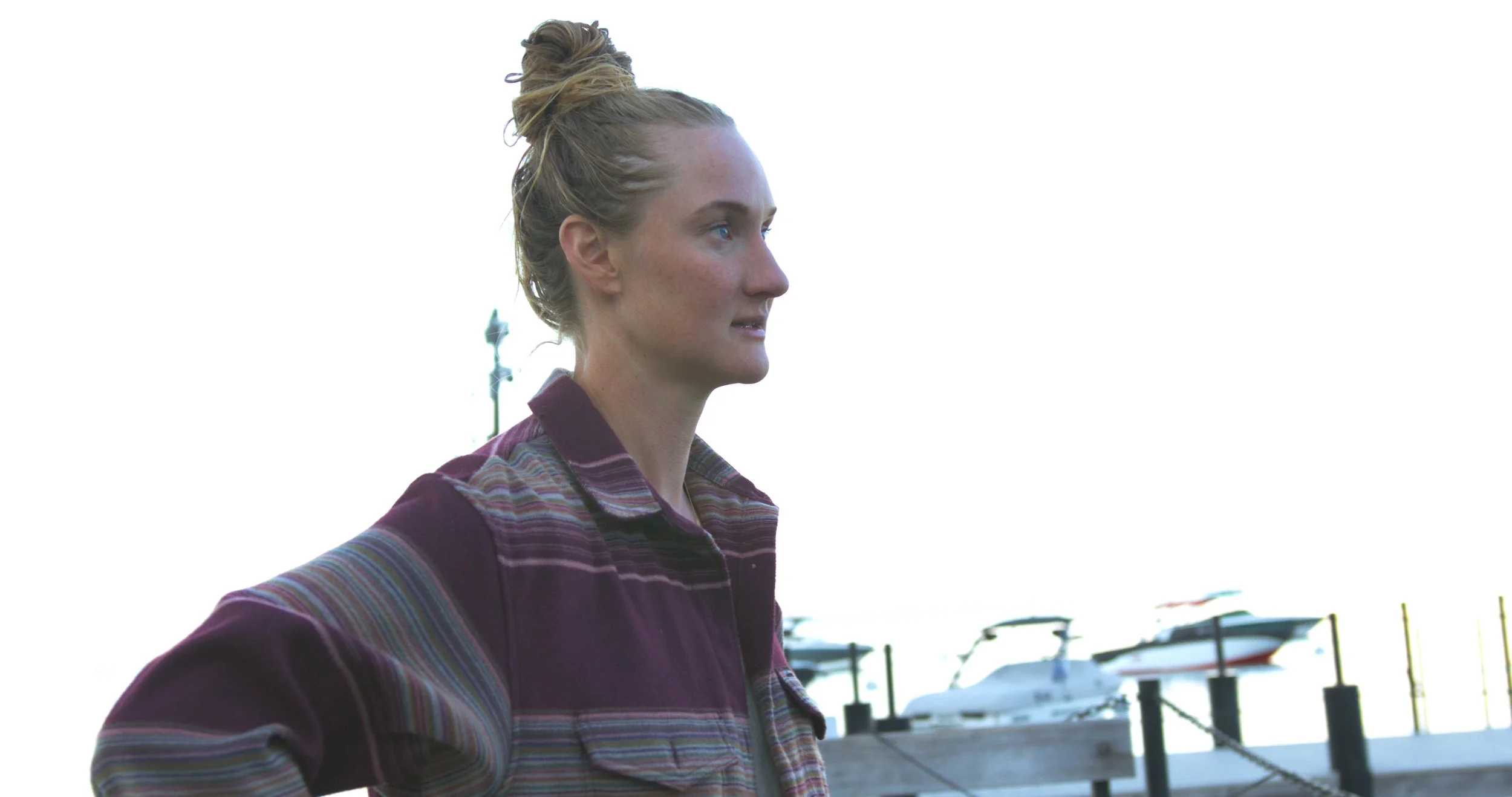 A woman with blonde hair in a bun standing at a marina, wearing a plaid shirt with boats in the background.