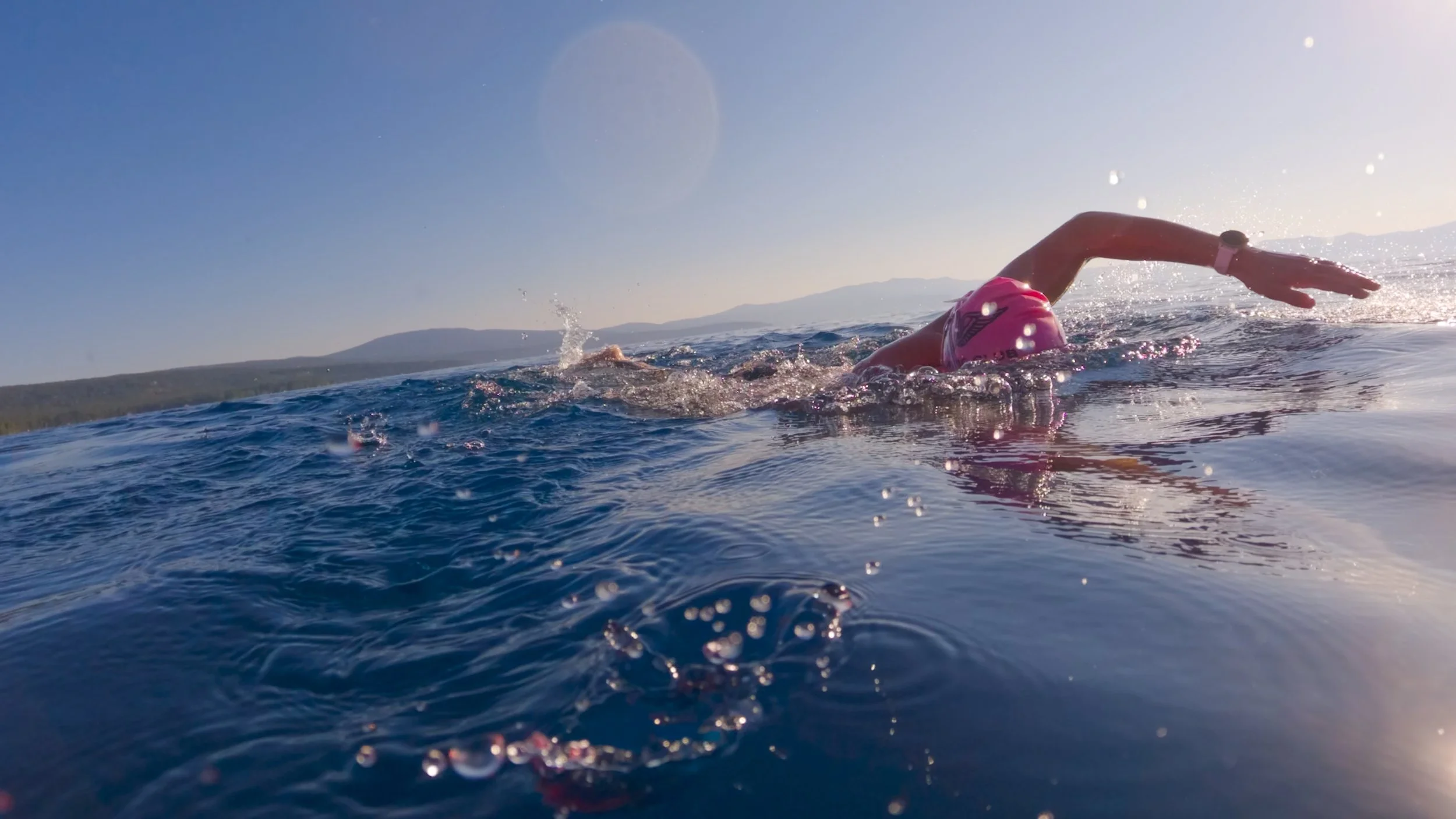 A swimmer in pink cap and wetsuit swimming in open water with mountains in the background during sunset.