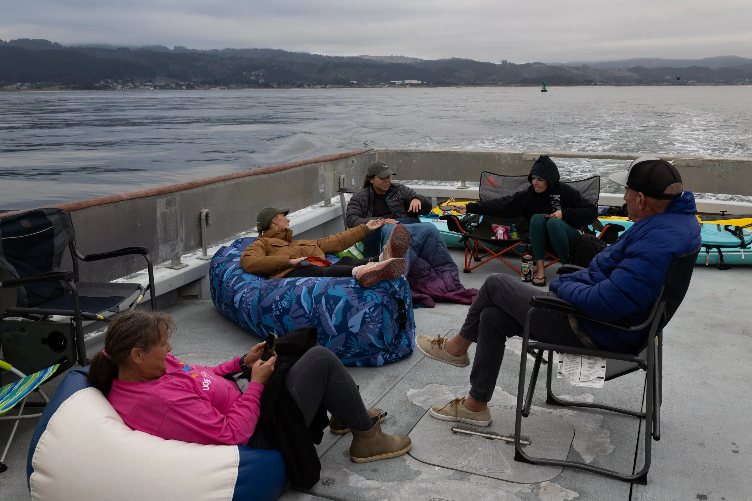 Group of five people relaxing on a boat deck, some seated or lying on chairs and cushions, with kayak paddles and water visible in the background under cloudy skies.