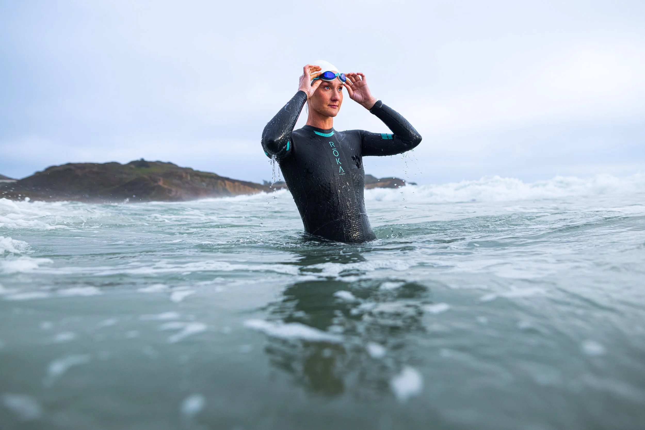 A woman wearing a black wetsuit and goggles stands in the ocean, adjusting her goggles with a mountainous island in the background.