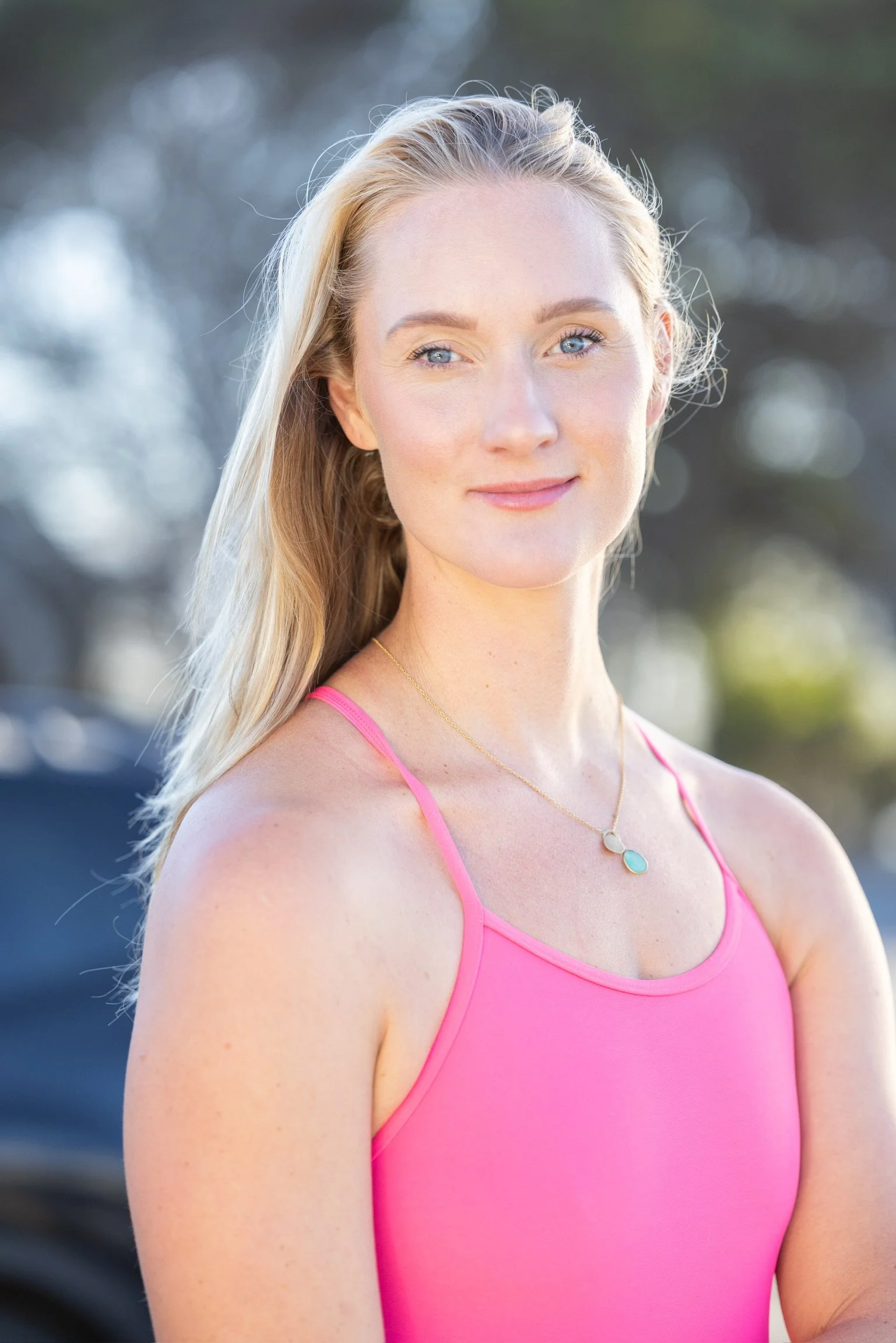 A young woman with blonde hair and blue eyes, wearing a pink tank top and a delicate necklace, outdoors with blurred trees in the background.