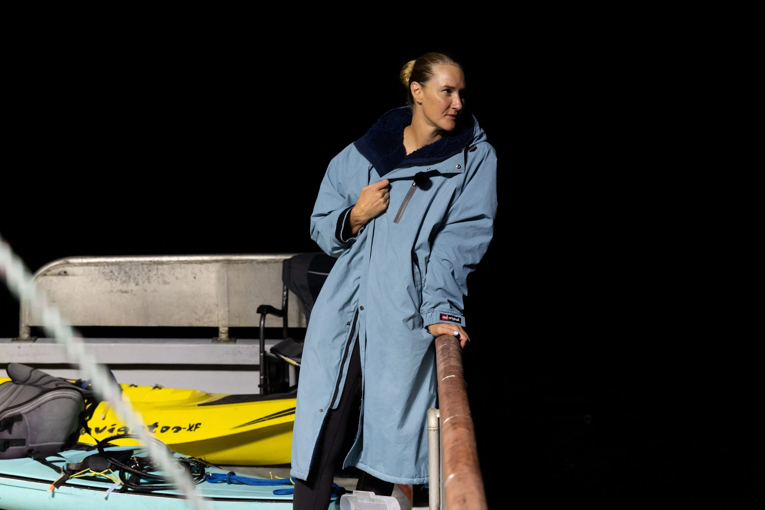 A woman in a blue coat leaning on a boat railing at night, with boats and gear visible in the background.