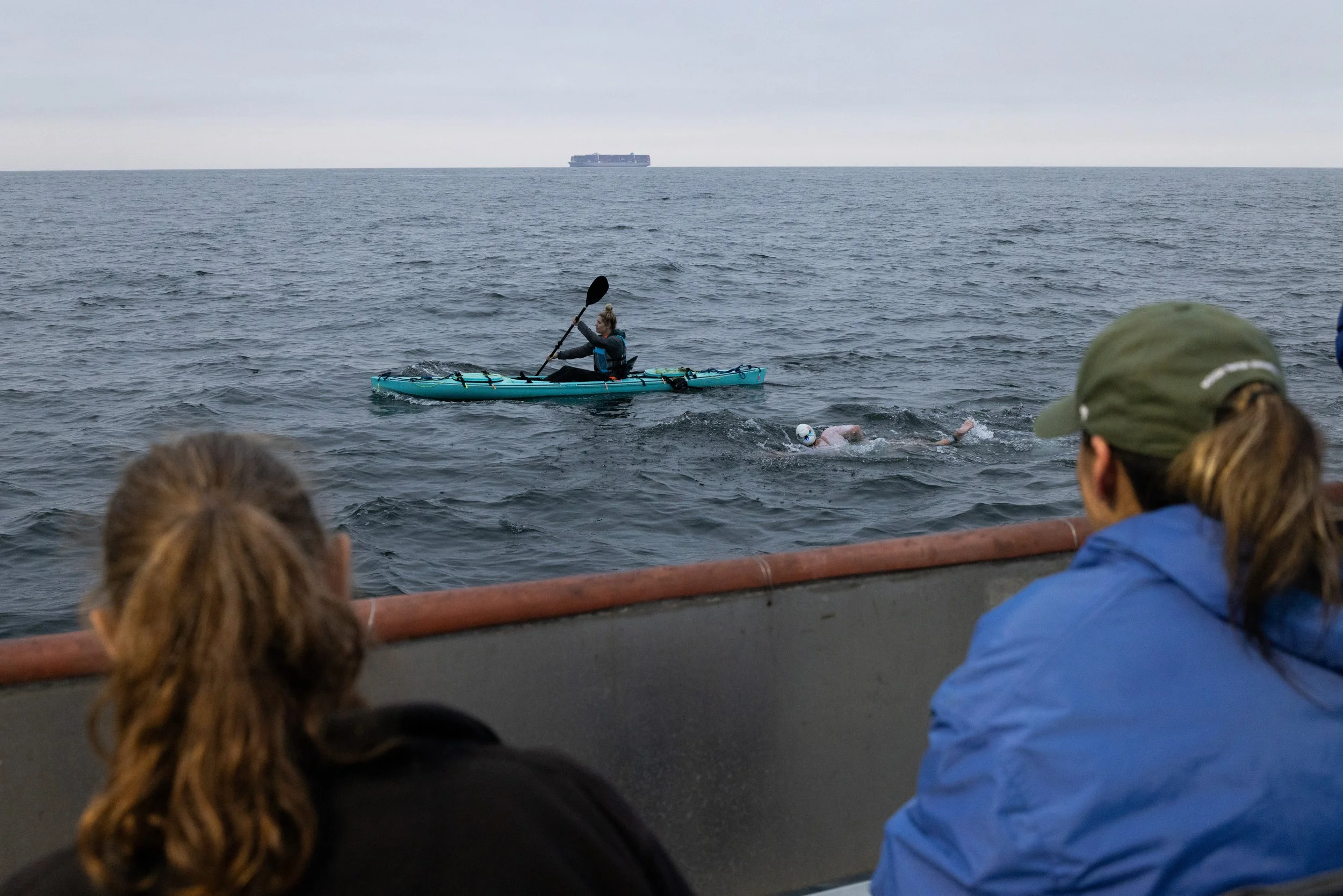 People observing a swimmer in the ocean, with a kayak nearby and a large cargo ship in the distance on overcast day.