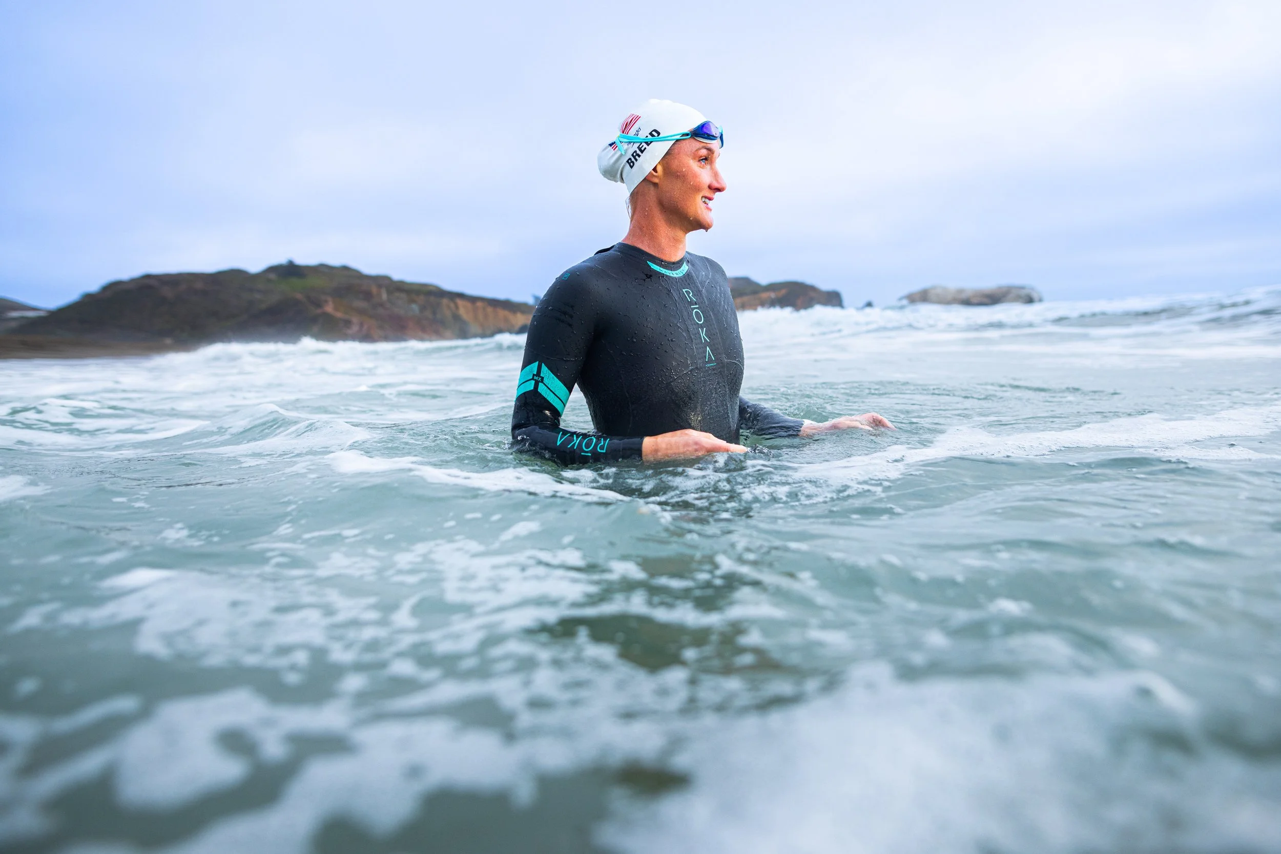 Woman in black wetsuit and white swim cap standing in ocean water during daytime with hills and cloudy sky in background.