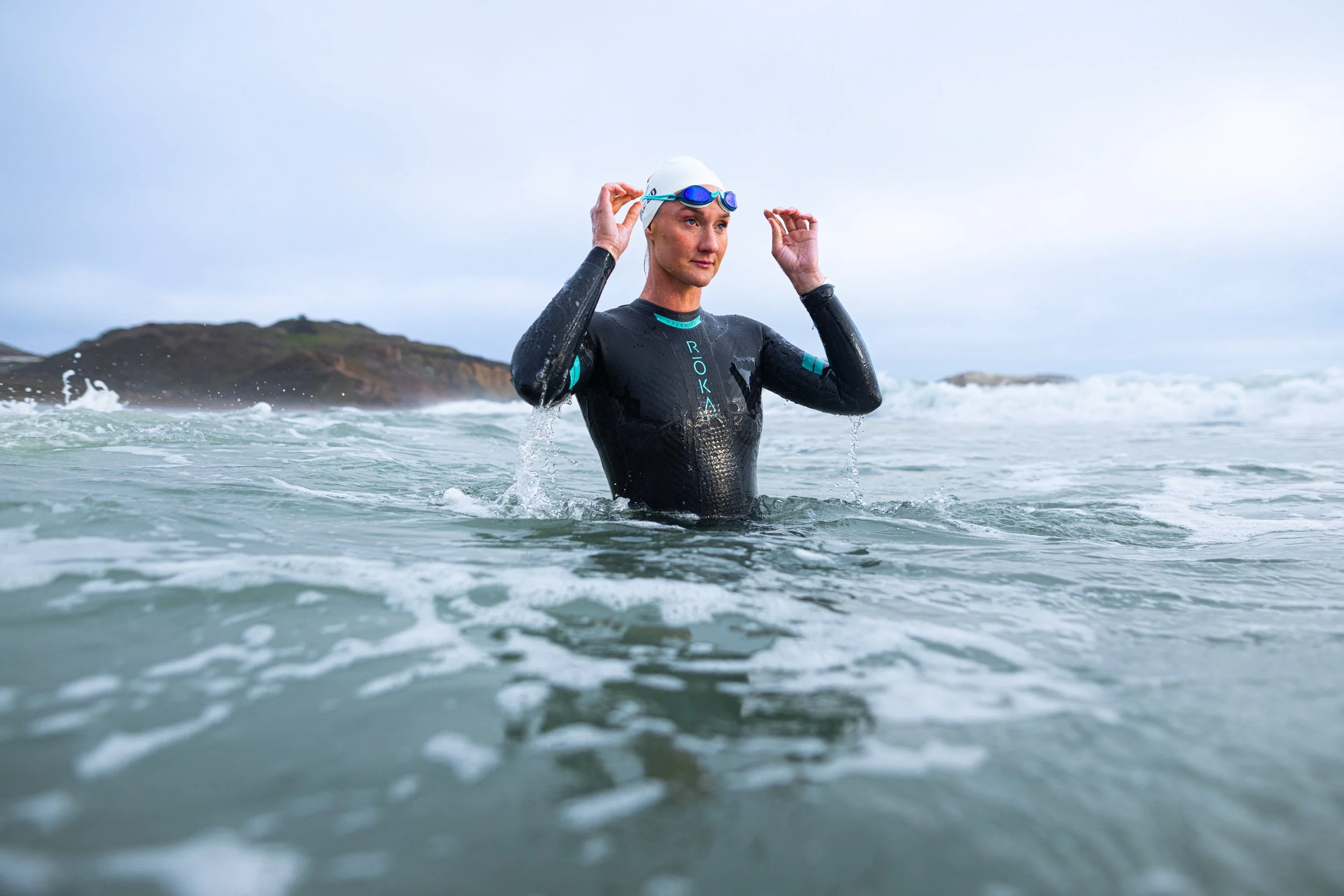 Woman in wetsuit and swimming cap adjusting goggles in the ocean, with waves and an island in the background.